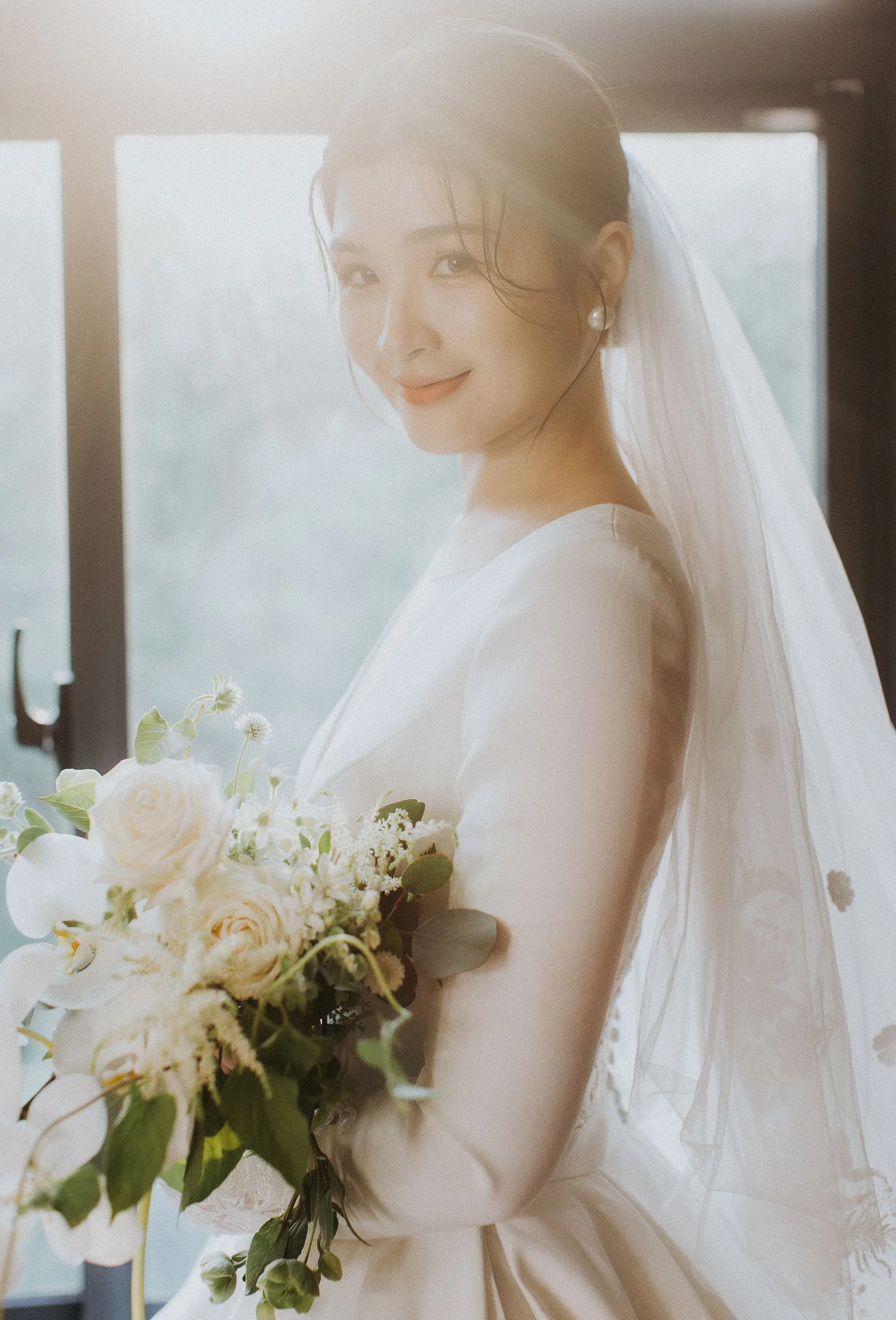A woman in a wedding dress holds a bouquet of white and cream flowers with greenery, smiling with sunlight streaming in from a large window behind her.
