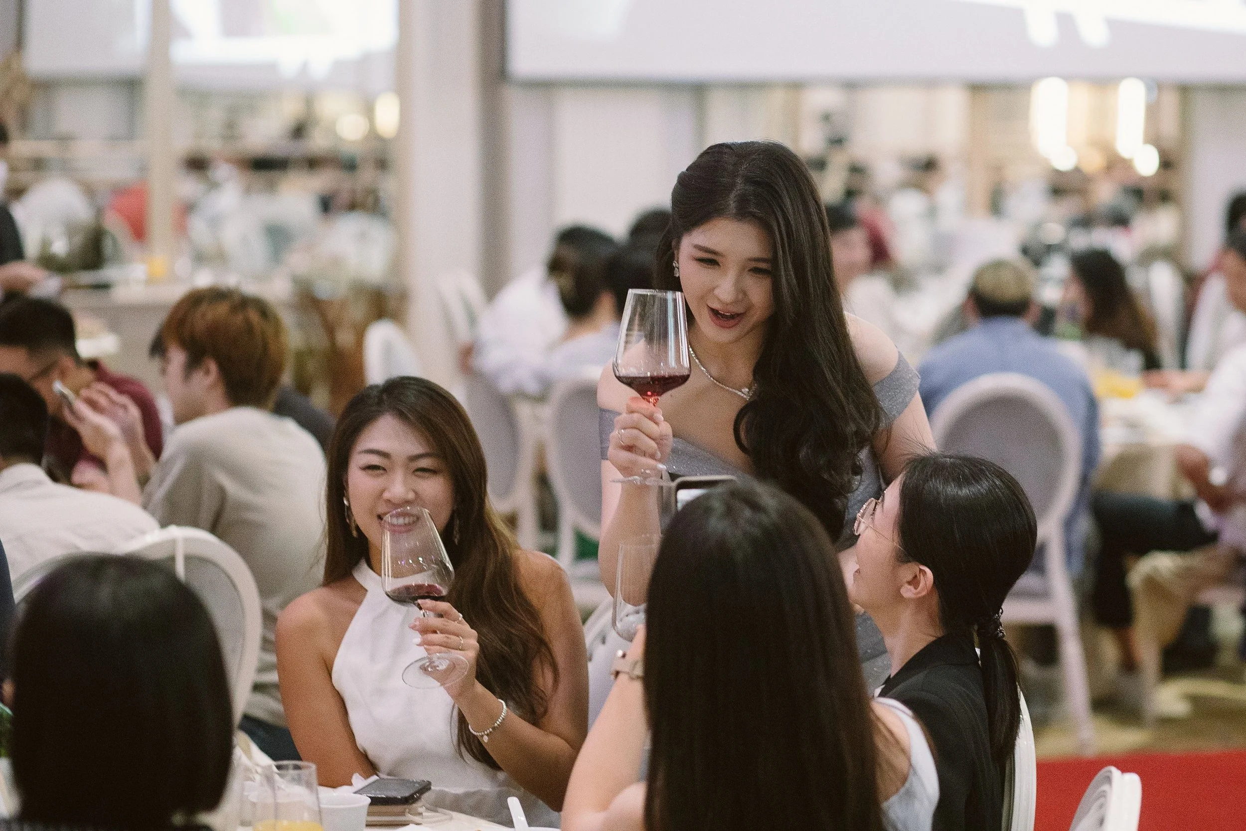 A woman in a gray dress with long dark hair smiling as she holds a glass of red wine at a social gathering in a brightly lit indoor setting with many people seated at tables in the background.