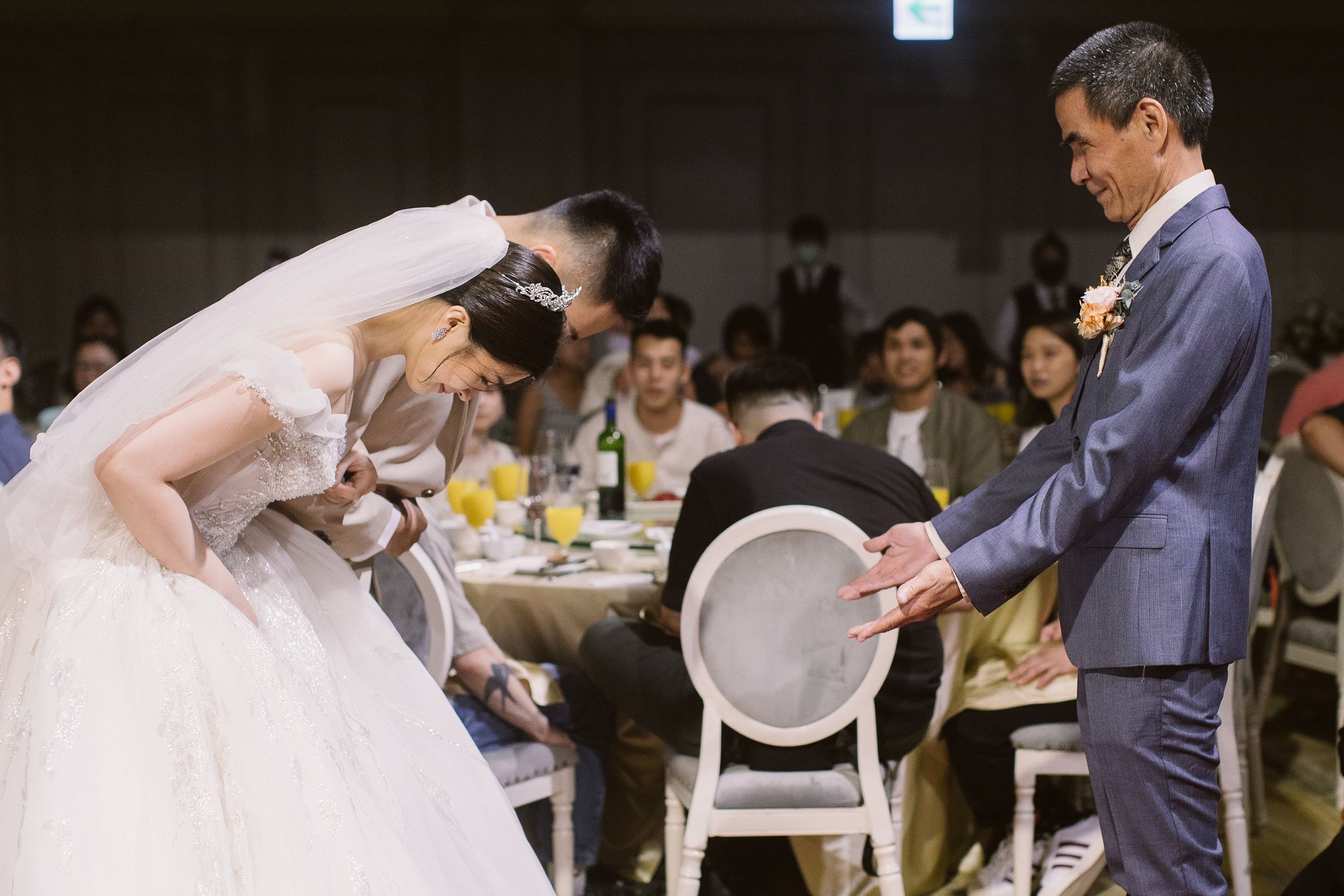 A bride and groom bow to their wedding guests during a wedding ceremony, with guests seated at tables in the background.