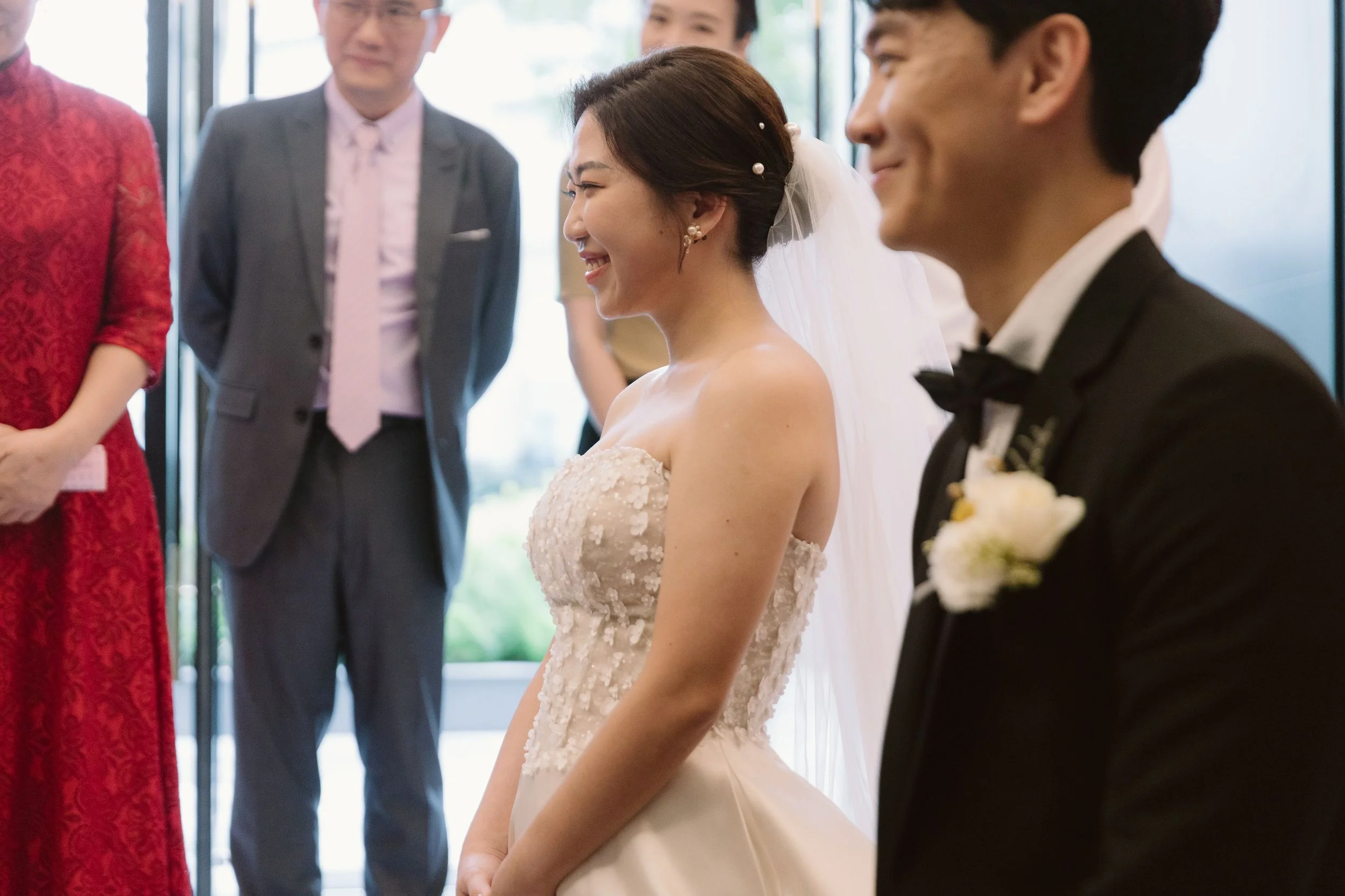A bride in a strapless white wedding gown smiling with her eyes closed during a wedding ceremony, surrounded by guests including a man in a tuxedo and others in formal attire.