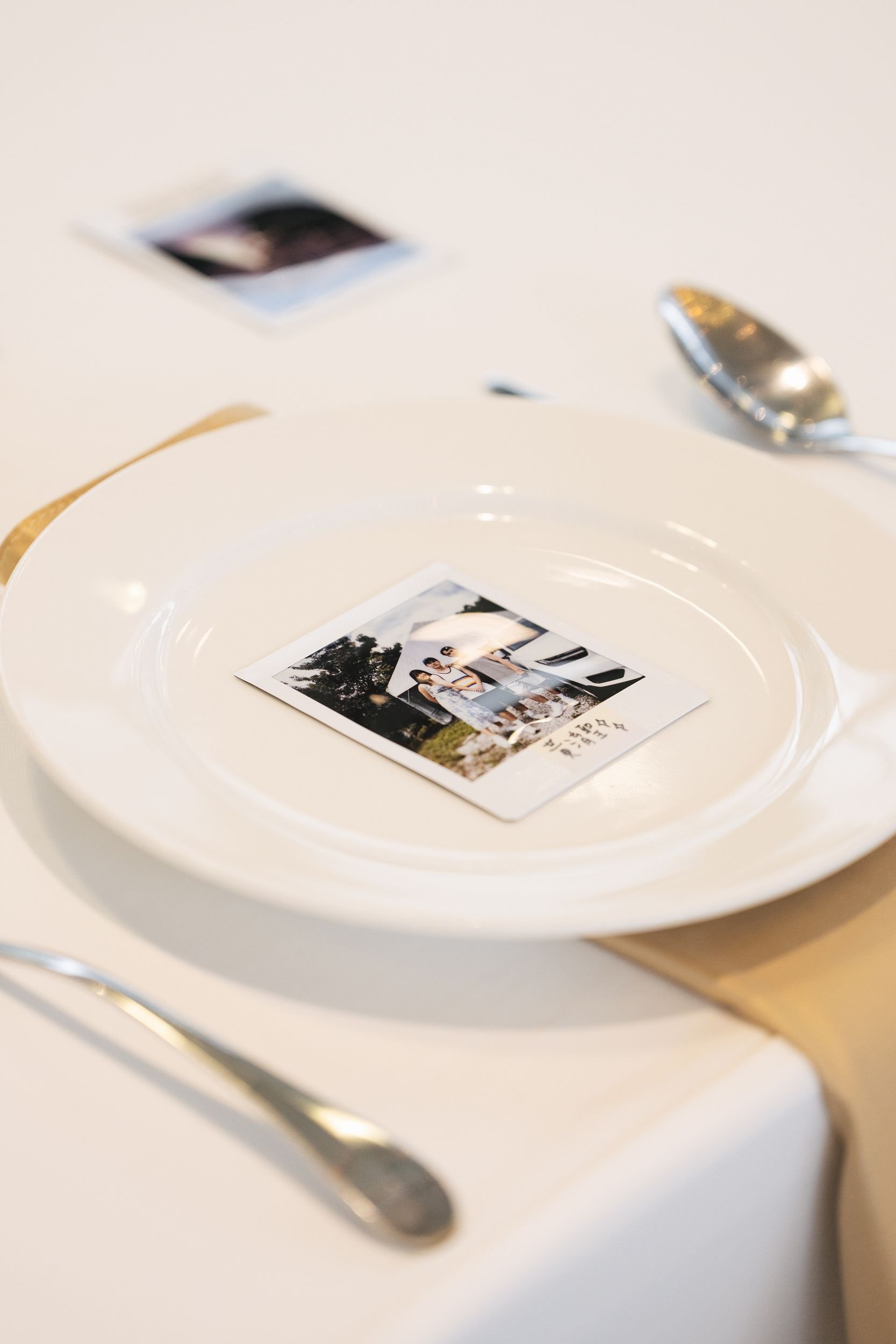 A photograph of a white plate with a small Polaroid photo of two women outdoors, on a table with a spoon and silverware, in a well-lit setting.