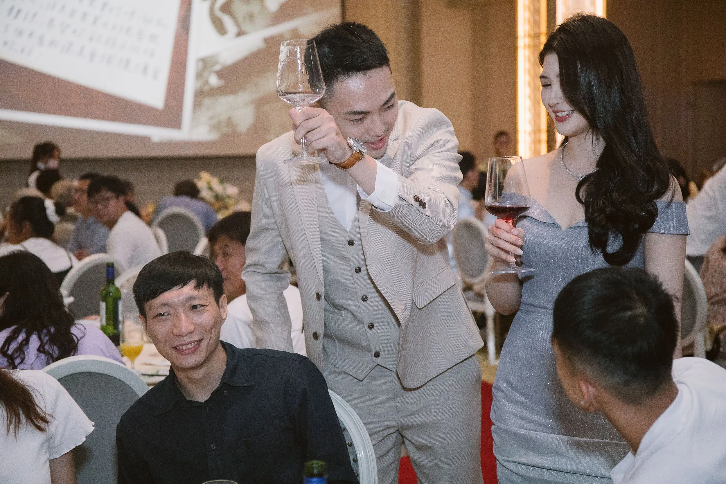 A man and a woman are smiling and conversing at a formal event, holding glasses of red wine. The man is wearing a light-colored suit, and the woman is in a shimmering silver dress. There are other guests seated at tables in the background.