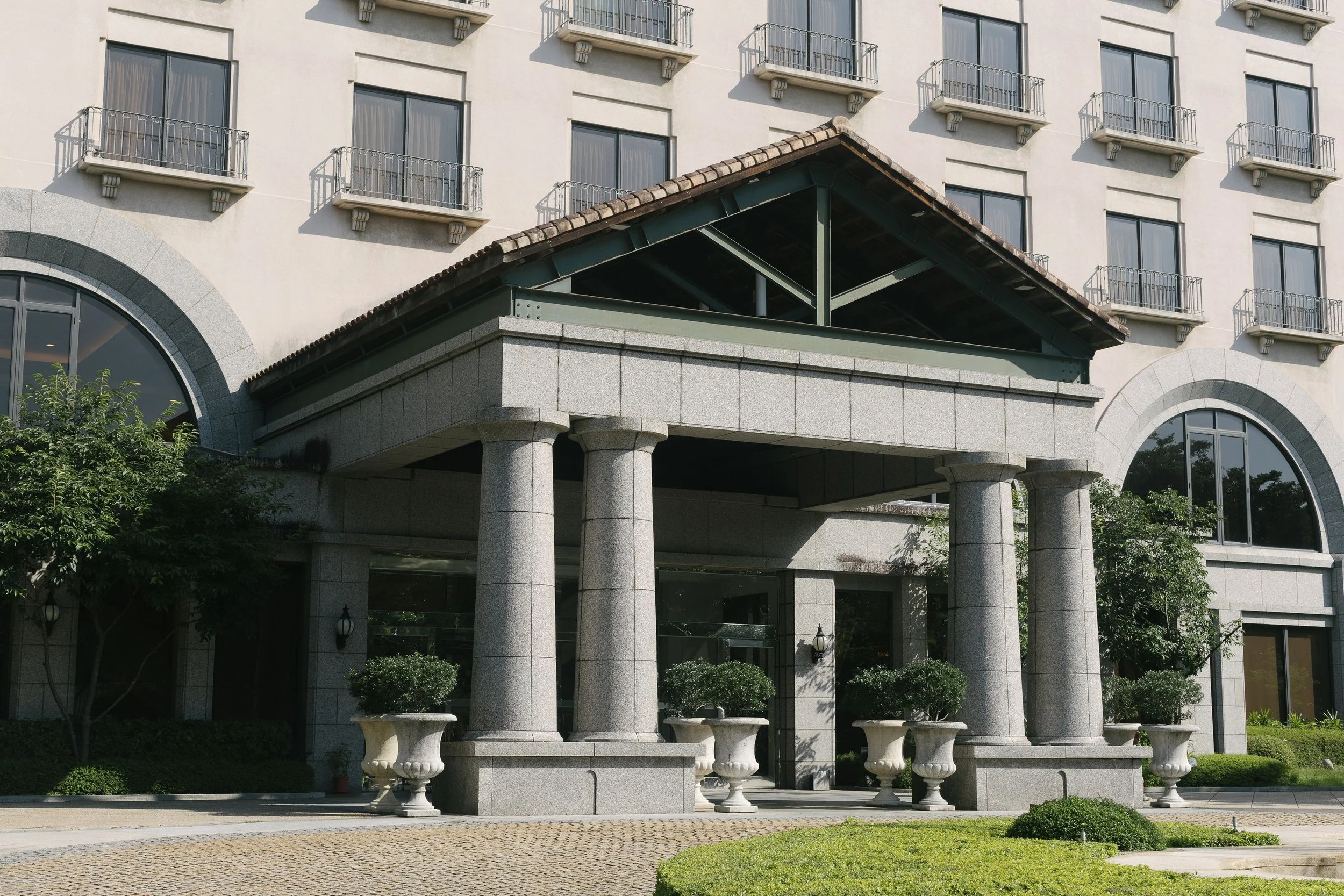 Facade of a multi-story building with windows and balconies. The entrance area has large stone columns, a covered porch with a tile roof, and outdoor seating benches. Surrounding greenery includes trees and bushes.