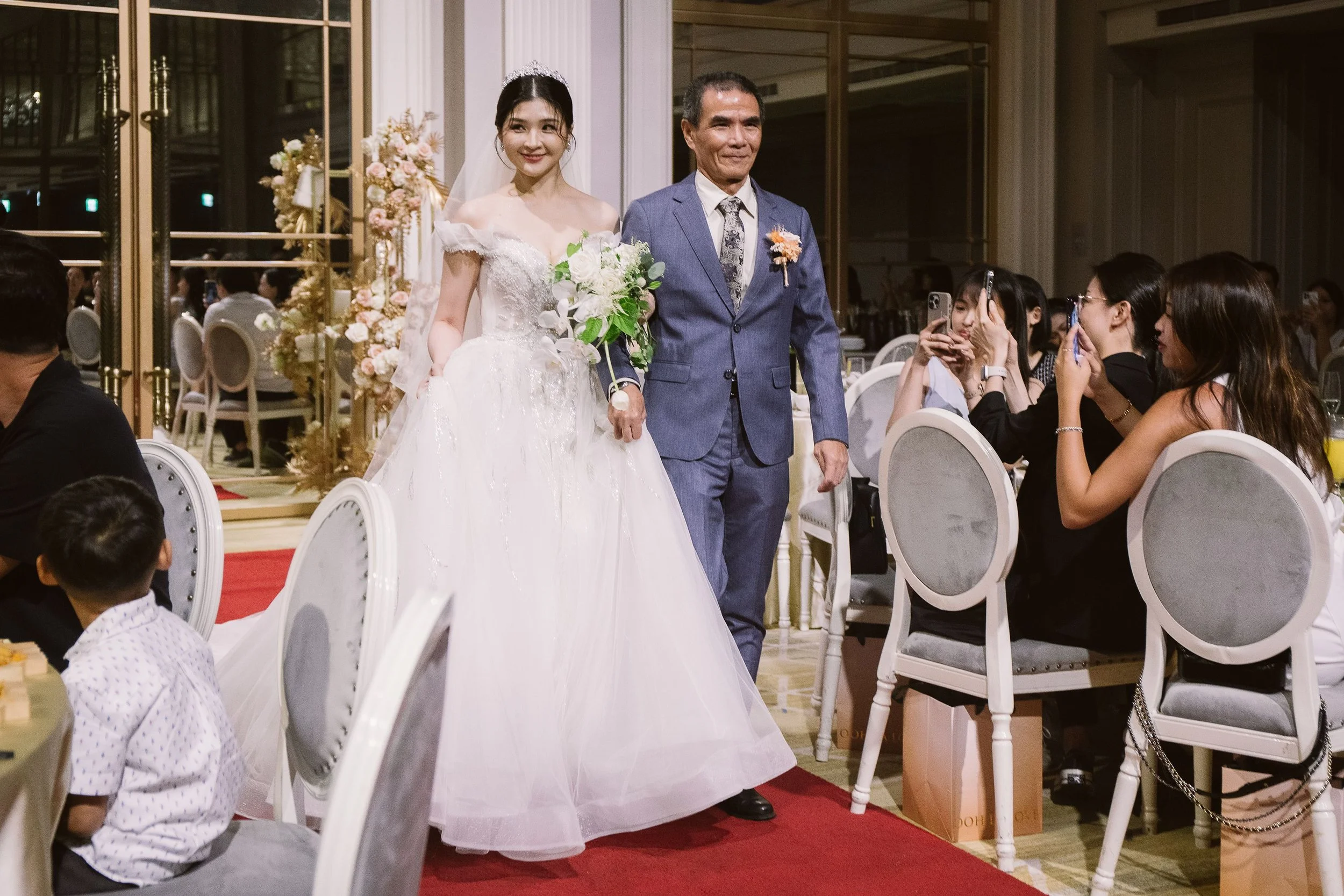 A bride in a white wedding gown holding a bouquet, walking down the aisle, flanked by seated guests taking photos, with a man in a blue suit walking beside her, in a decorated banquet hall.
