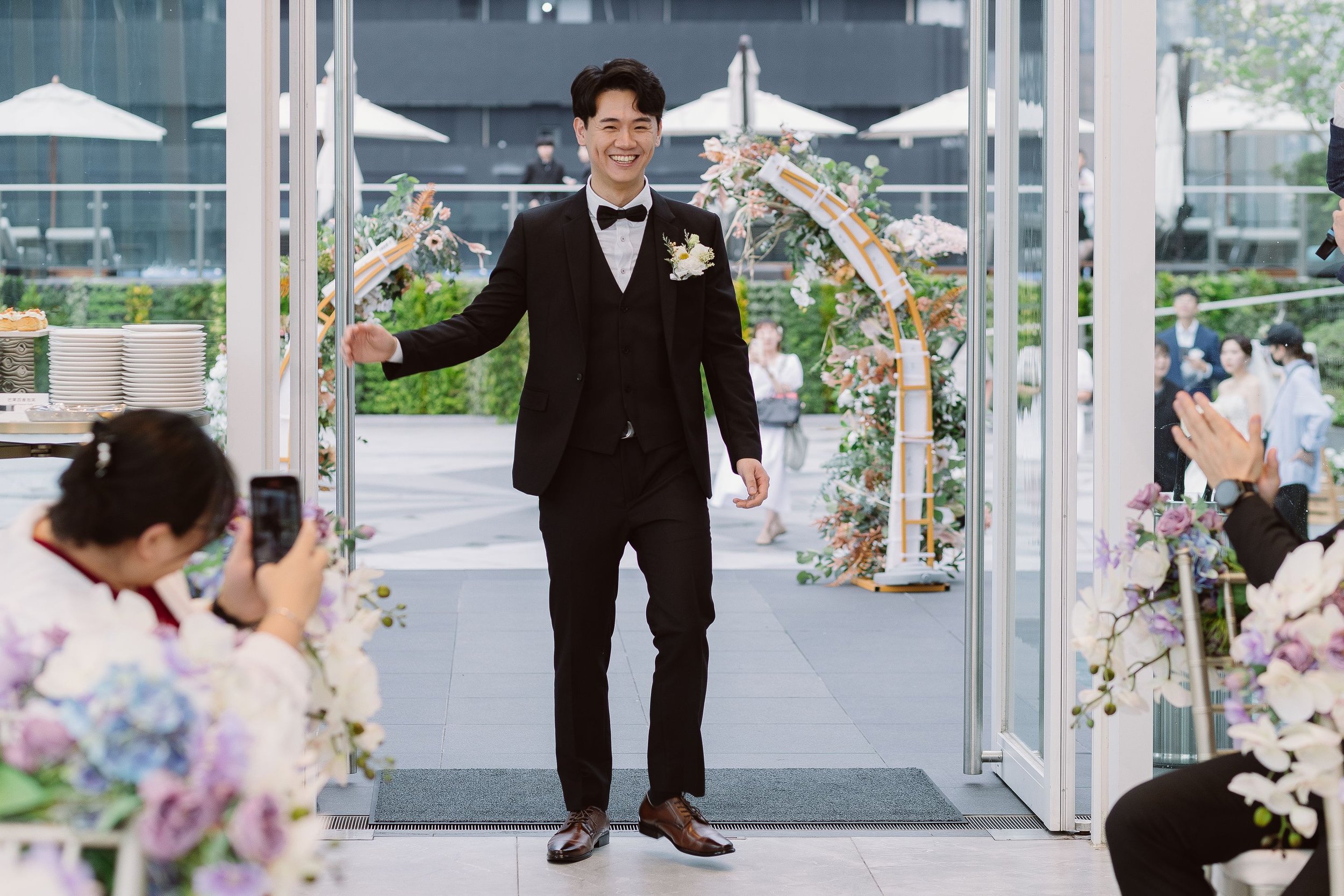 A man in a black suit and bow tie smiling as he walks through a doorway at a wedding or event, surrounded by floral decorations and seated guests.