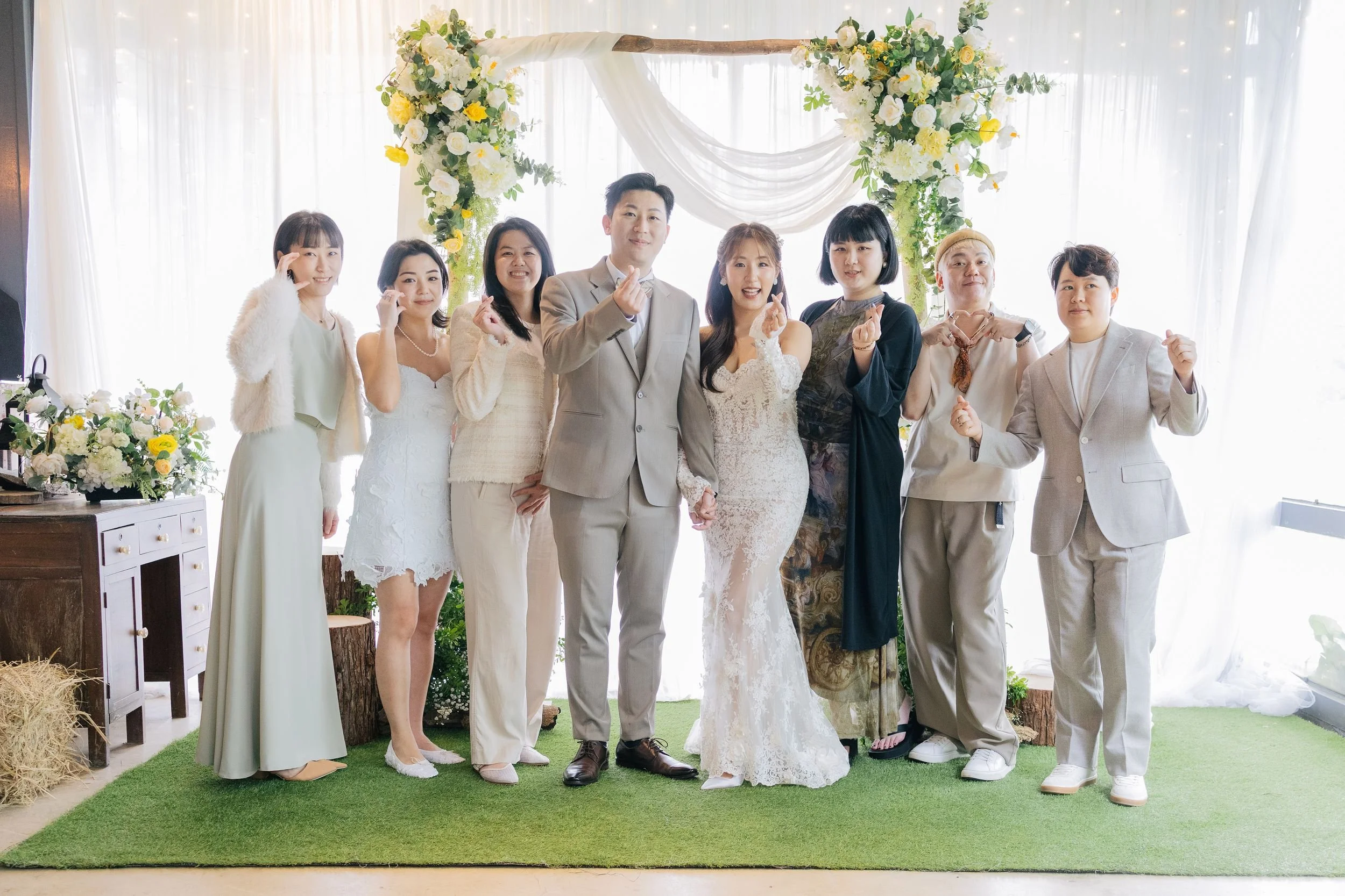 A group of seven people, including a bride and groom, posing in front of a wedding arch decorated with flowers and drapes at a wedding reception.