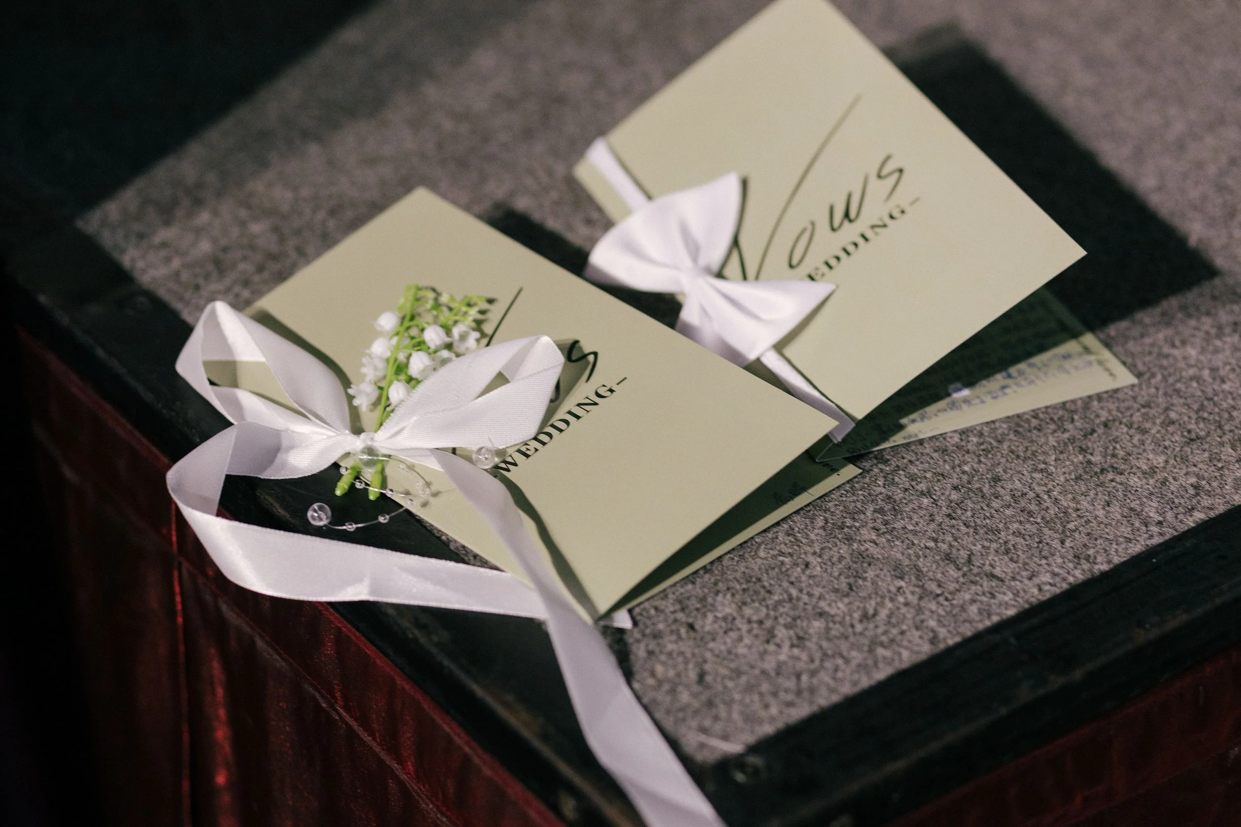 Wedding invitation cards decorated with white ribbons, small white flowers, and pearl accents are placed on a dark table.