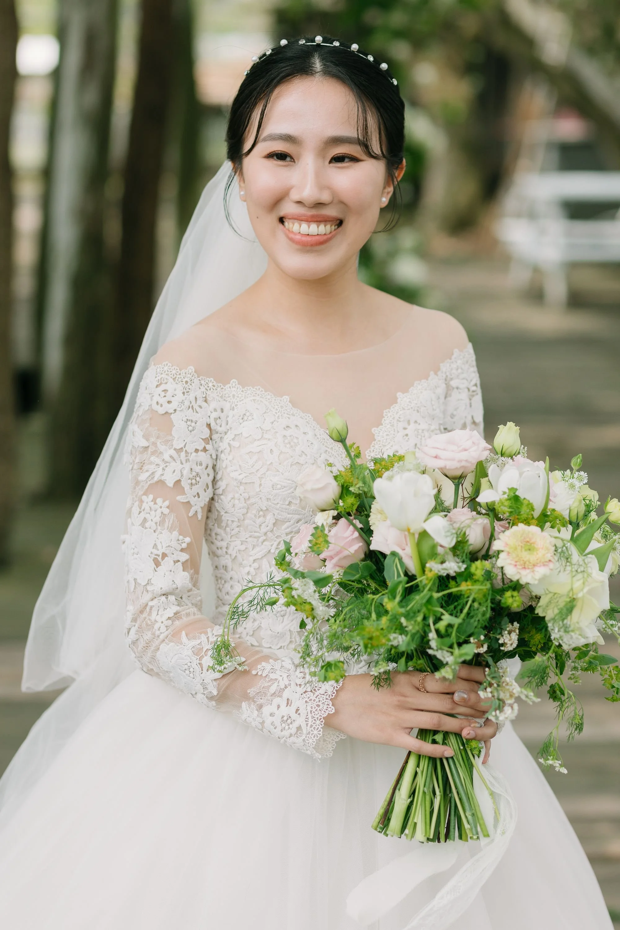 A smiling bride in a wedding dress holding a bouquet of pink and white flowers outdoors.