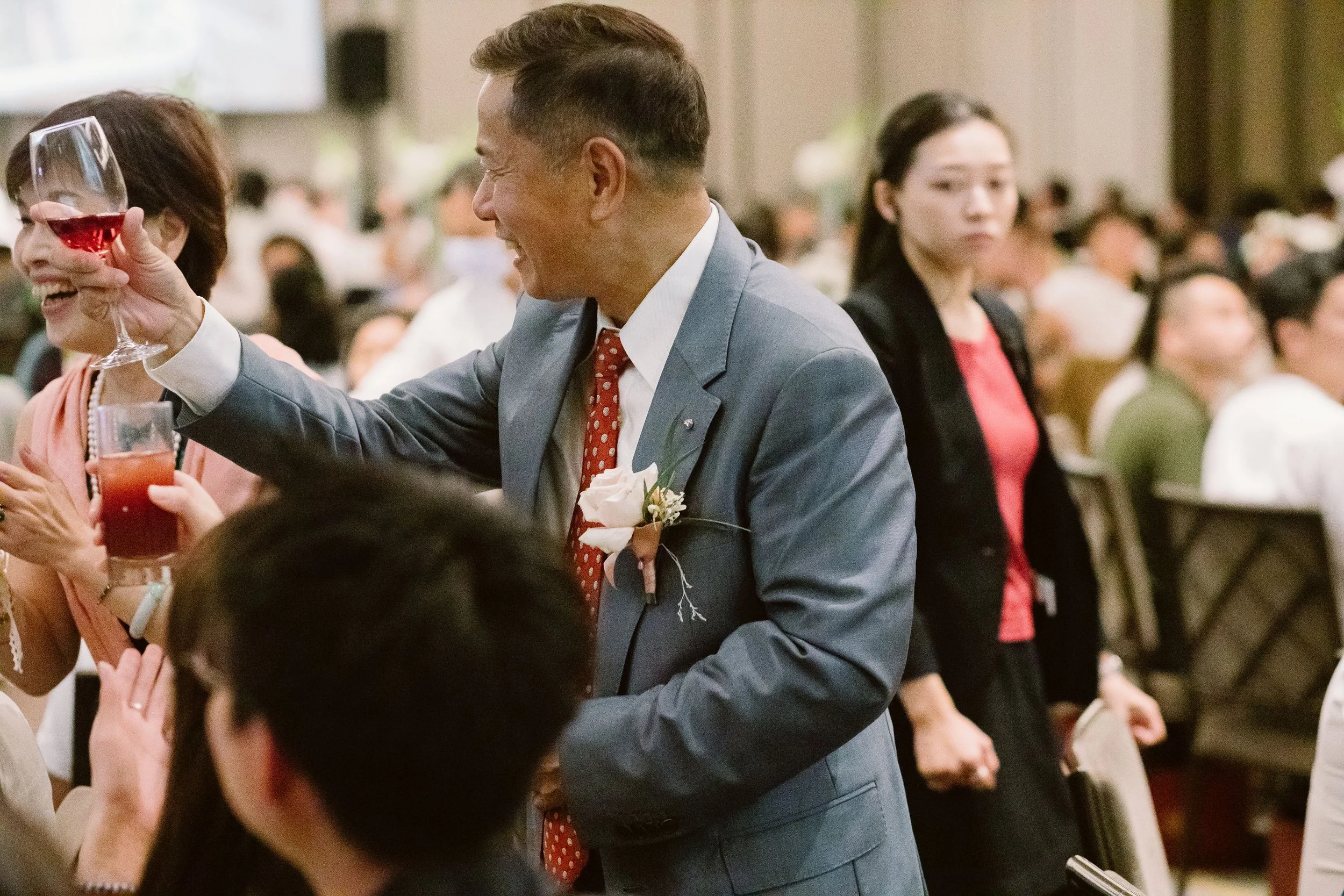 A man in a suit and tie with a flower pinned on his lapel, smiling and holding a glass of red wine at a social gathering. There are several people in the background, some sitting and some standing, in a large indoor event space.