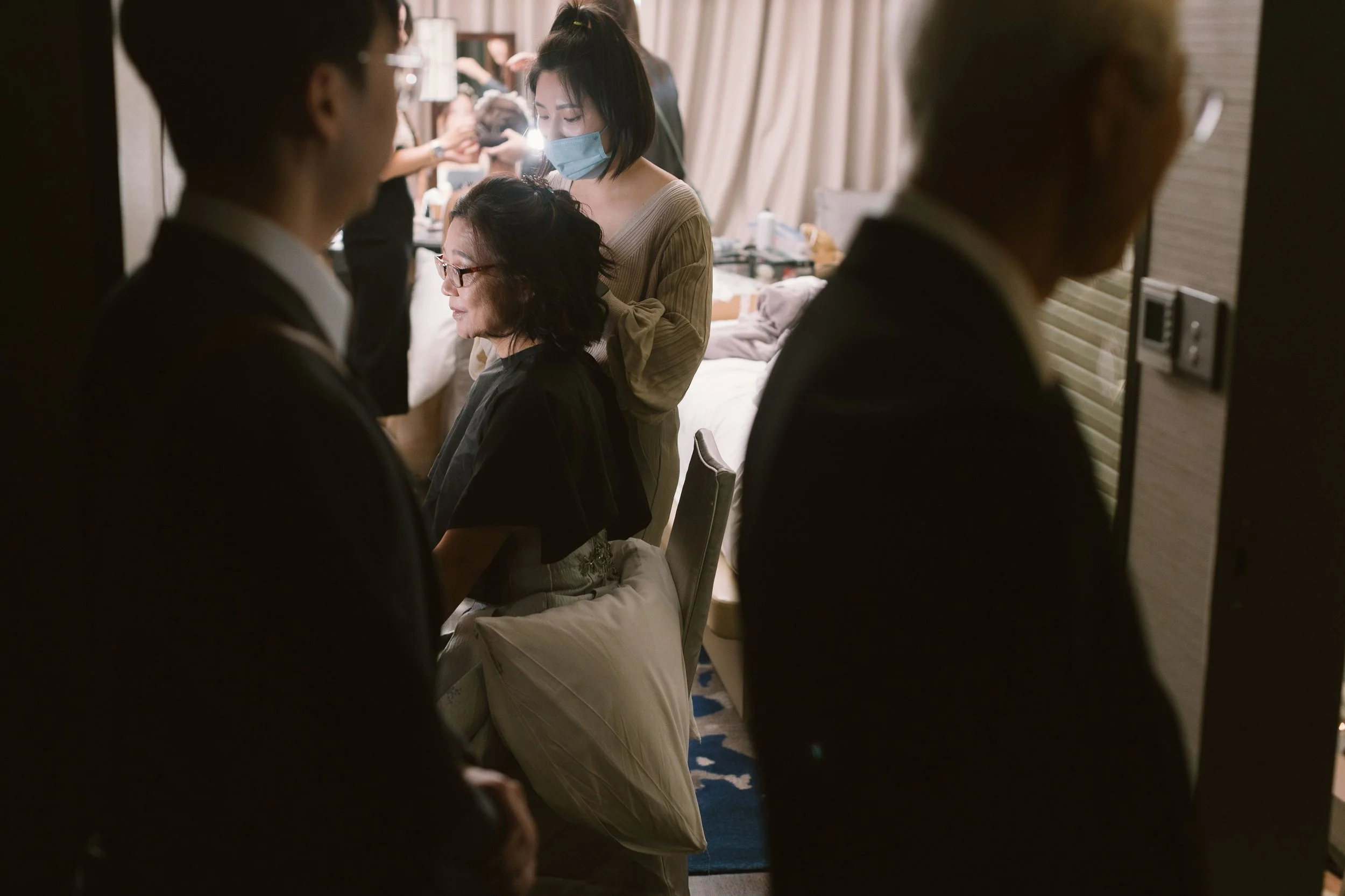 A woman wearing a face mask getting her hair done by a hairstylist in a clinic or salon setting, with other people and salon equipment in the background.
