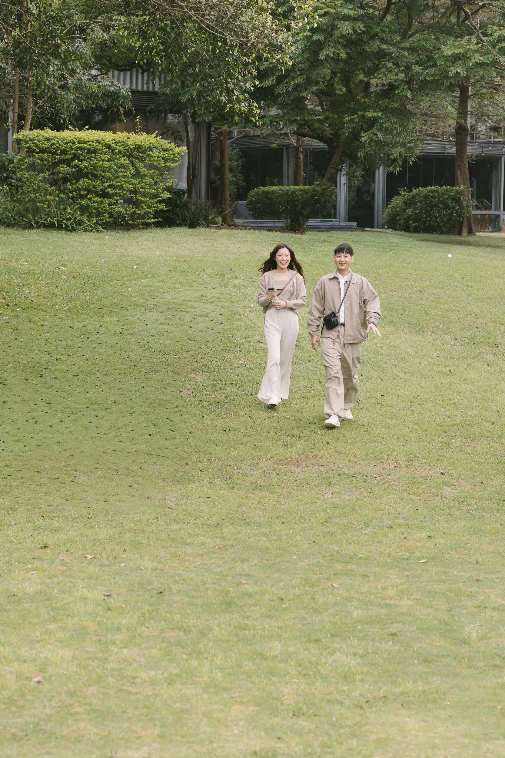 Two young people, a woman and a man, walking on a grassy lawn in a park with trees and bushes in the background. They are smiling and appear to be enjoying their time outdoors.