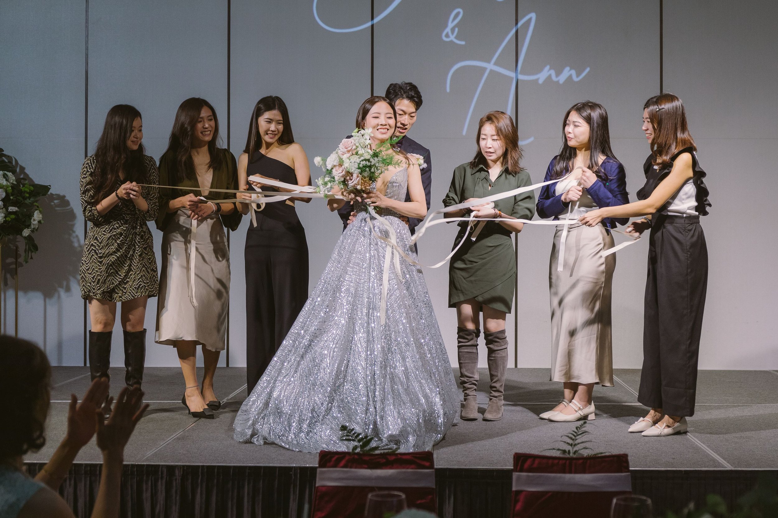 Group of women and a man standing on stage during a celebration or award ceremony. One woman in a silver dress is holding a bouquet of flowers, surrounded by smiling onlookers.
