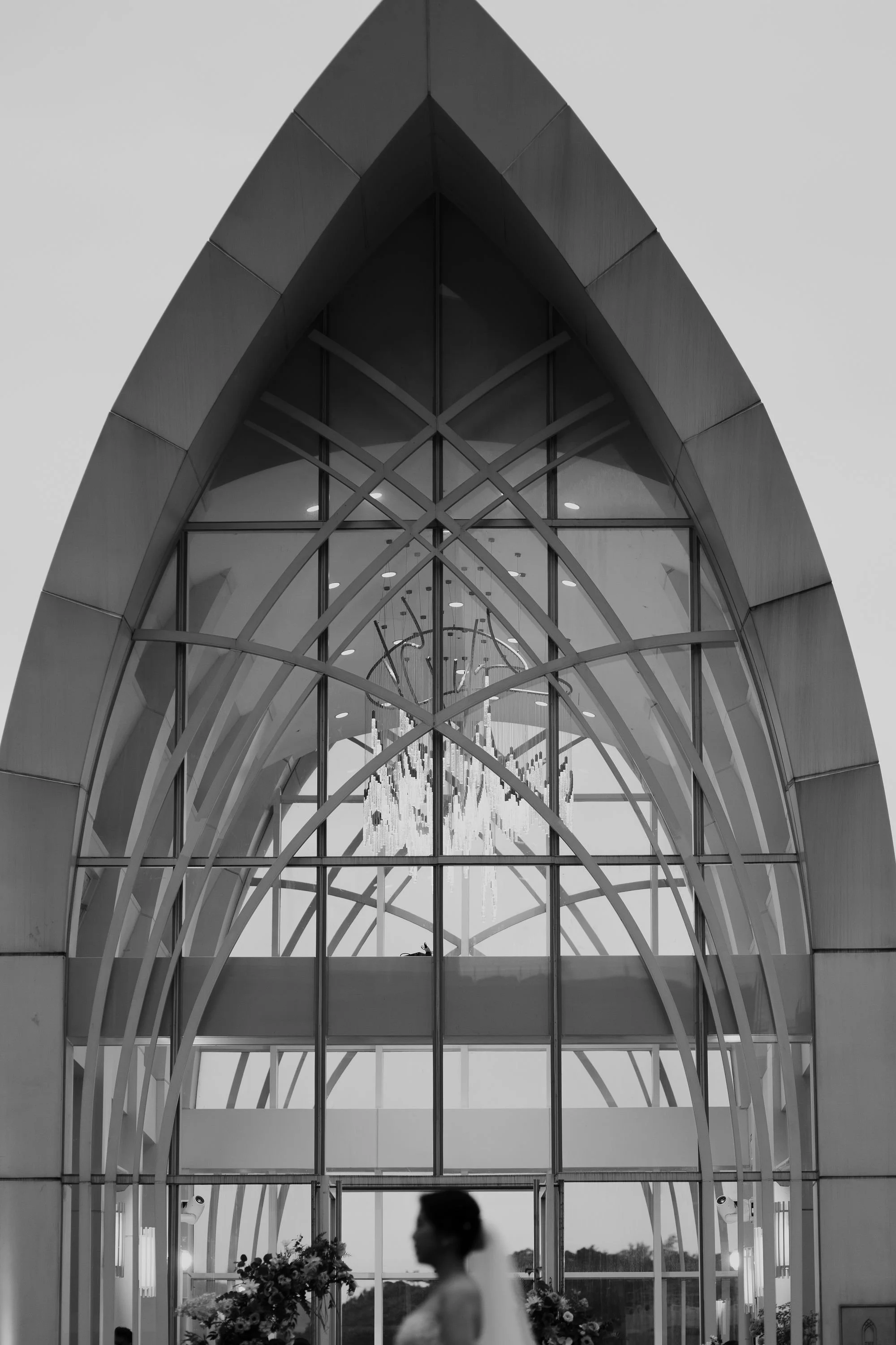 A grayscale photo of a modern architectural structure with a curved, glass-paneled roof and metal framework, with a person and floral arrangements visible at the bottom, possibly an event or ceremony taking place.