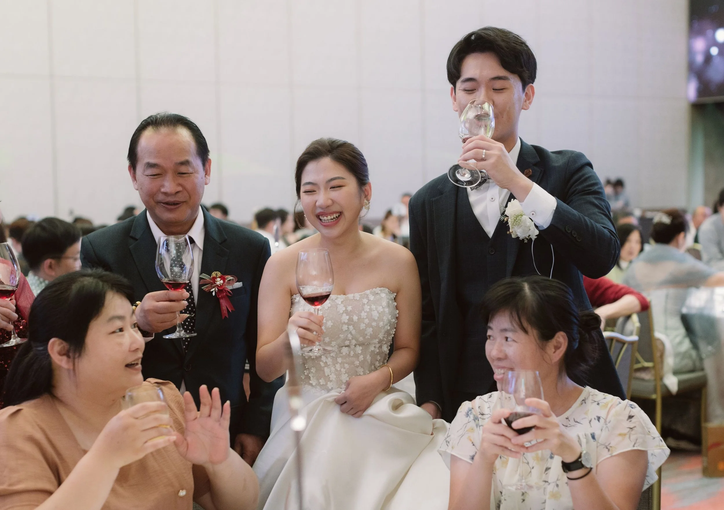People at a wedding reception enjoying drinks, with a bride in a white dress smiling in the center and a man in a suit and bow tie drinking from a wine glass nearby.