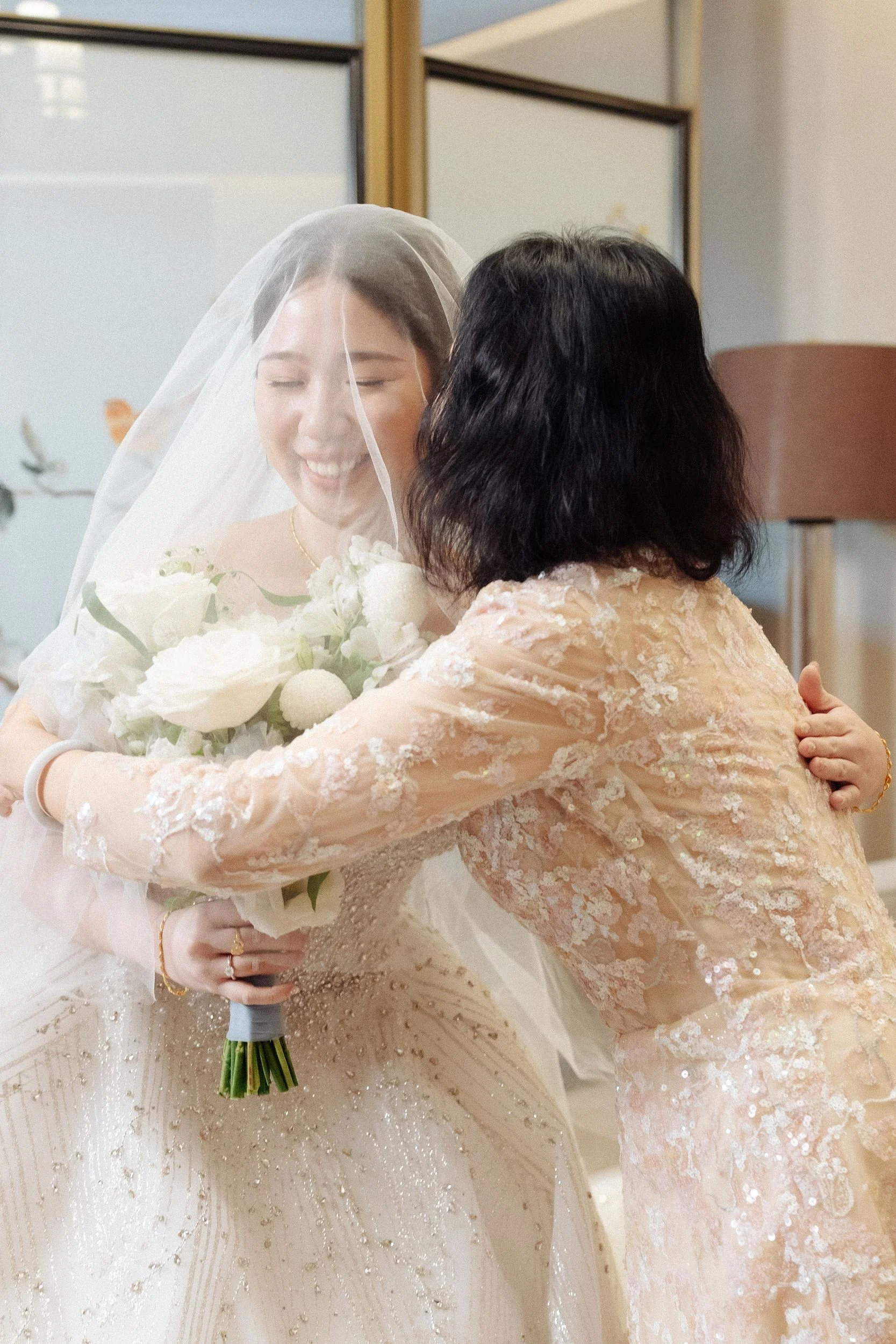 A bride with a veil holding a bouquet of white flowers embracing a woman with black hair dressed in a peach lace gown.