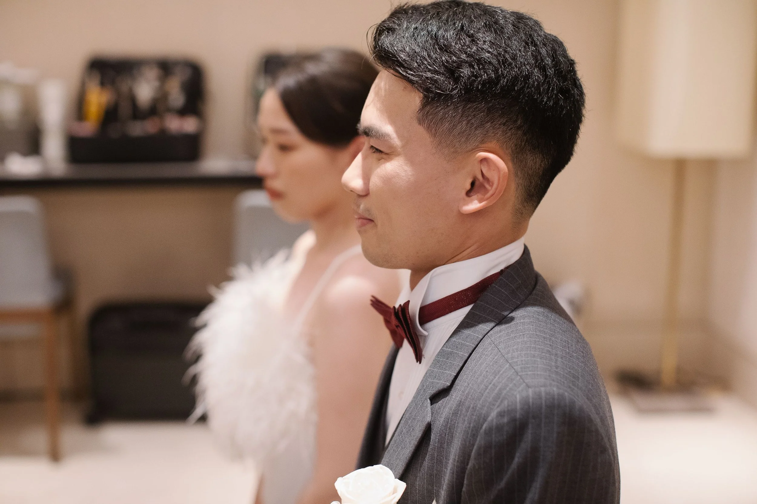 A man dressed in a gray suit and red bow tie sitting in a row beside a woman, both with eyes closed in a relaxed posture, possibly in a wedding or formal event setting.