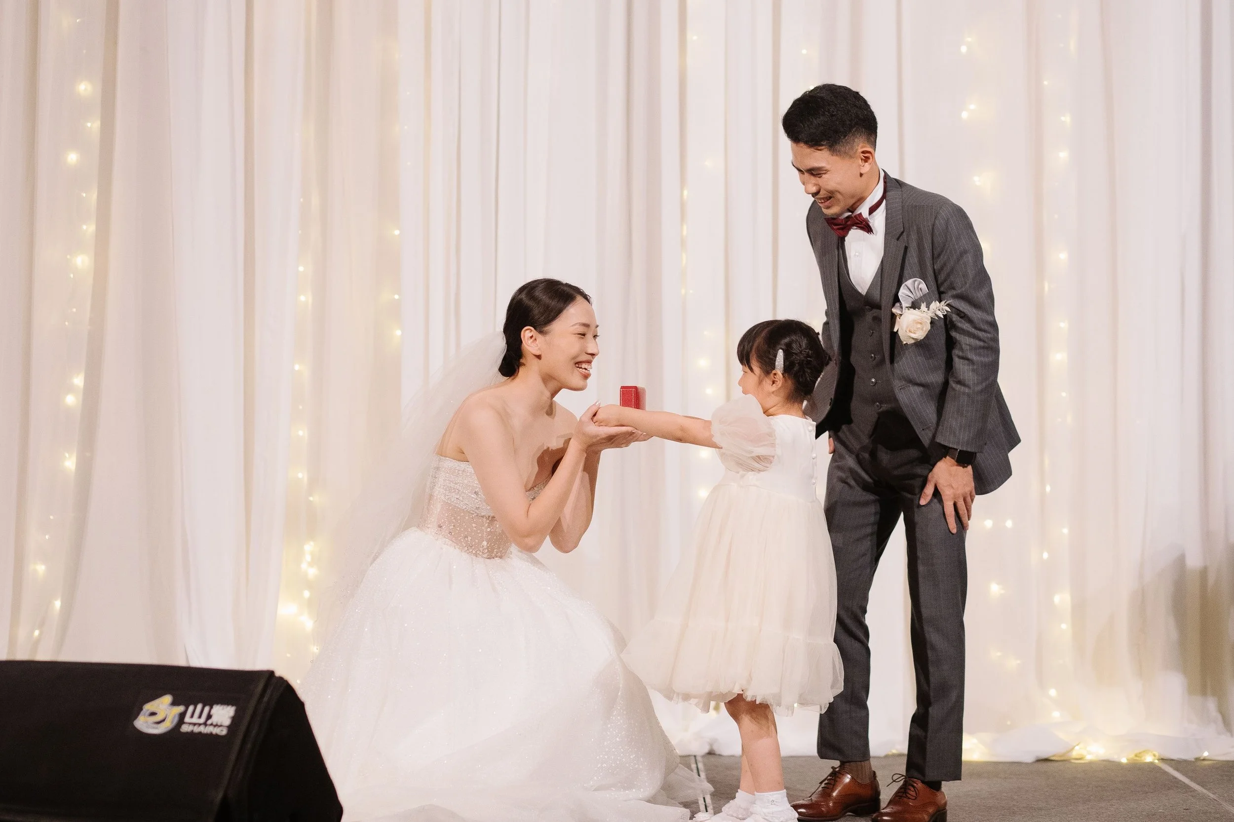 A wedding ceremony with a bride, groom, and a young girl exchanging rings in front of a backdrop of white curtains and string lights.