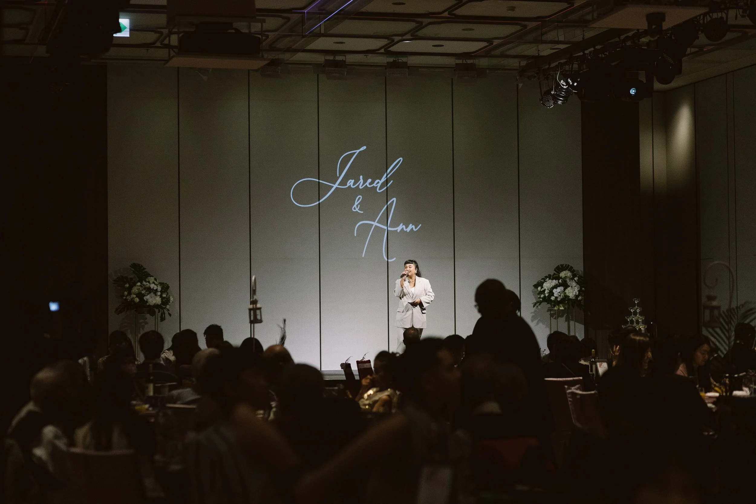 A woman in a white suit standing on a stage, speaking into a microphone, with the names Jared and Ann projected behind her. The setting appears to be a wedding or formal event with floral arrangements and a darkened audience in the foreground.