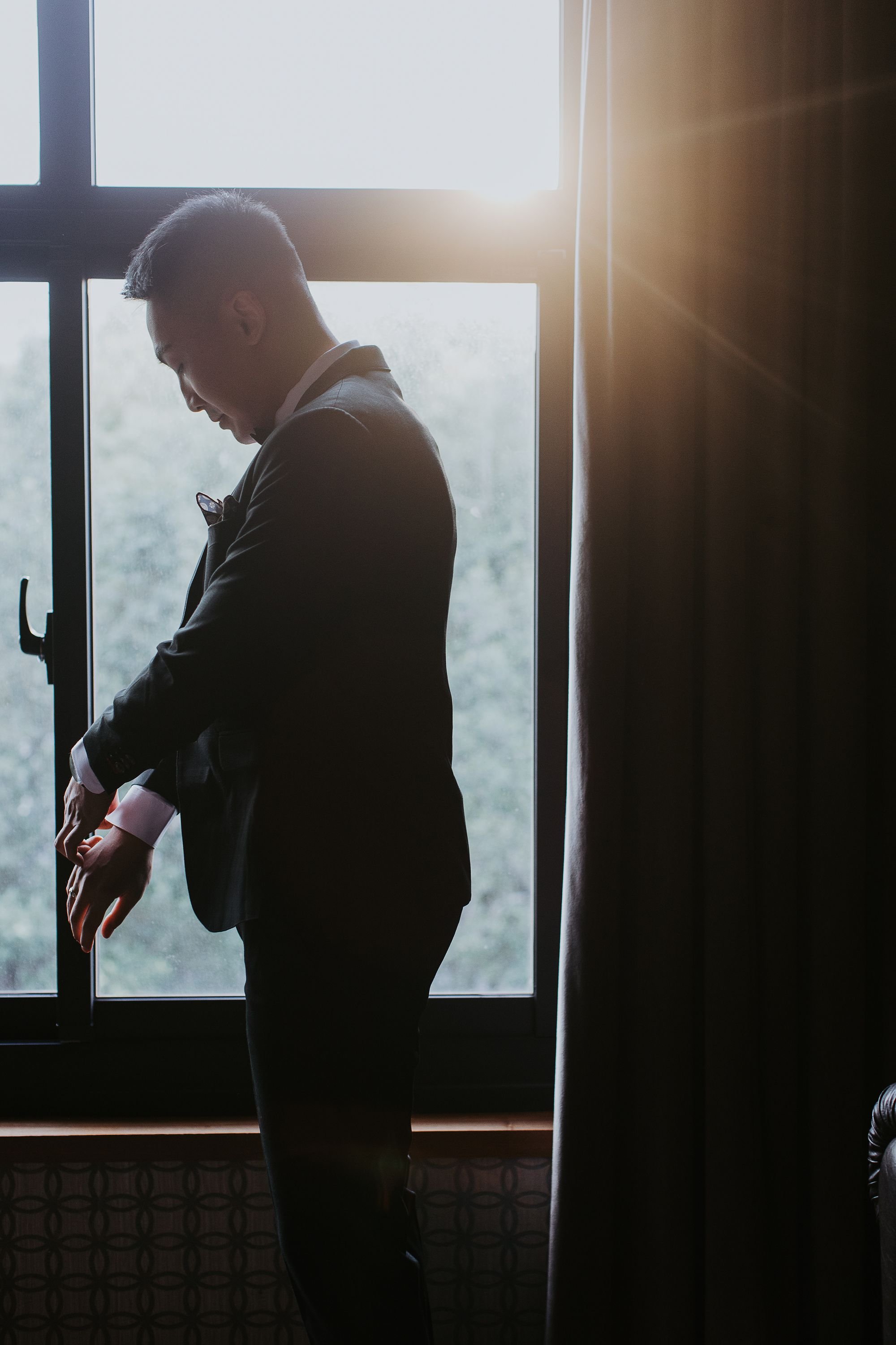 A man is standing indoors near a window with sunlight coming through. He is adjusting his cufflinks and appears to be getting dressed, probably for a formal occasion.