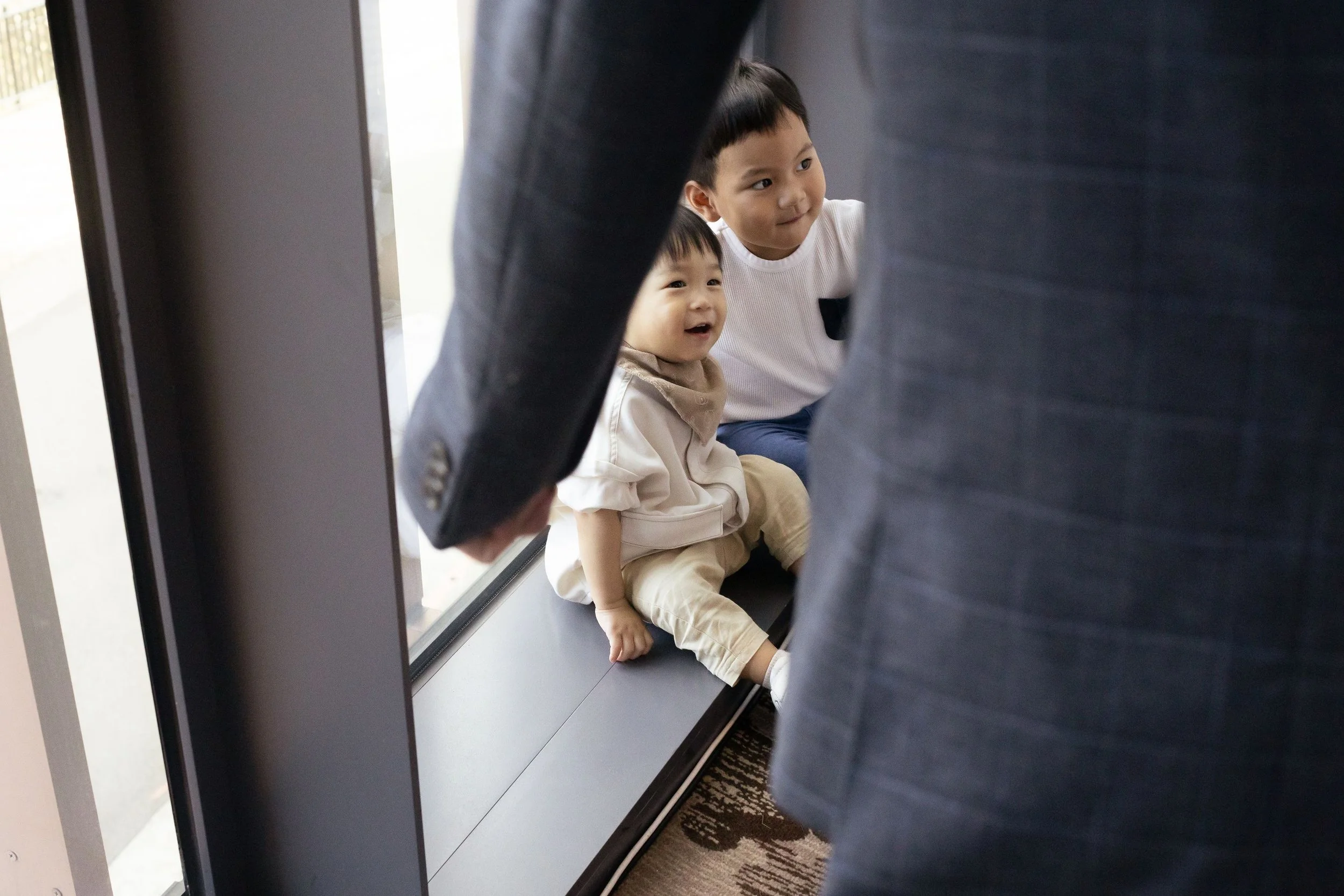 Two young boys sitting on the floor near a glass door, seen from behind through a gap between two adults wearing dark pants.
