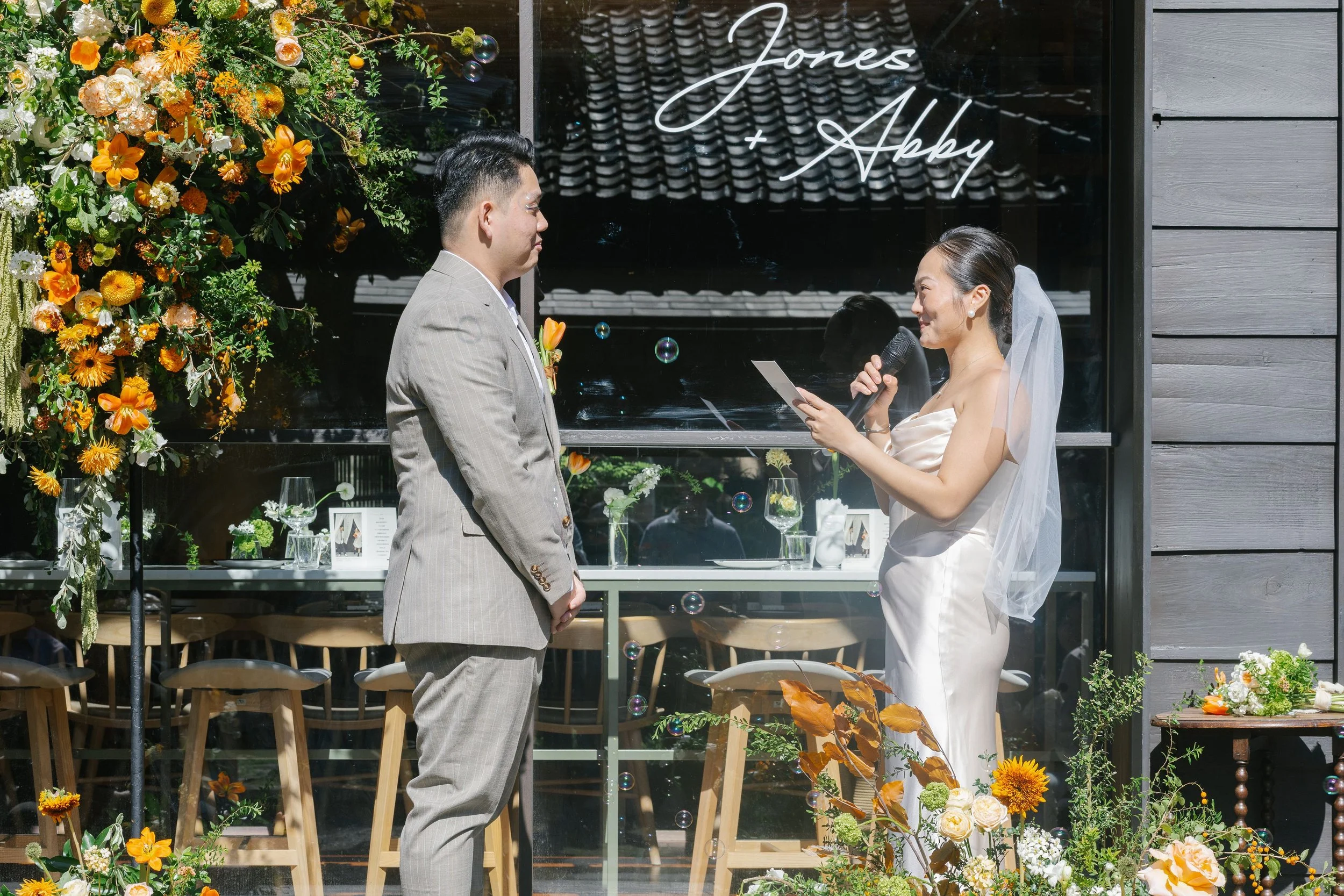 A bride and groom facing each other in front of a decorated backdrop with flowers and glasses, during a wedding ceremony outside a venue with signage 'Jones + Abby' in neon lights.