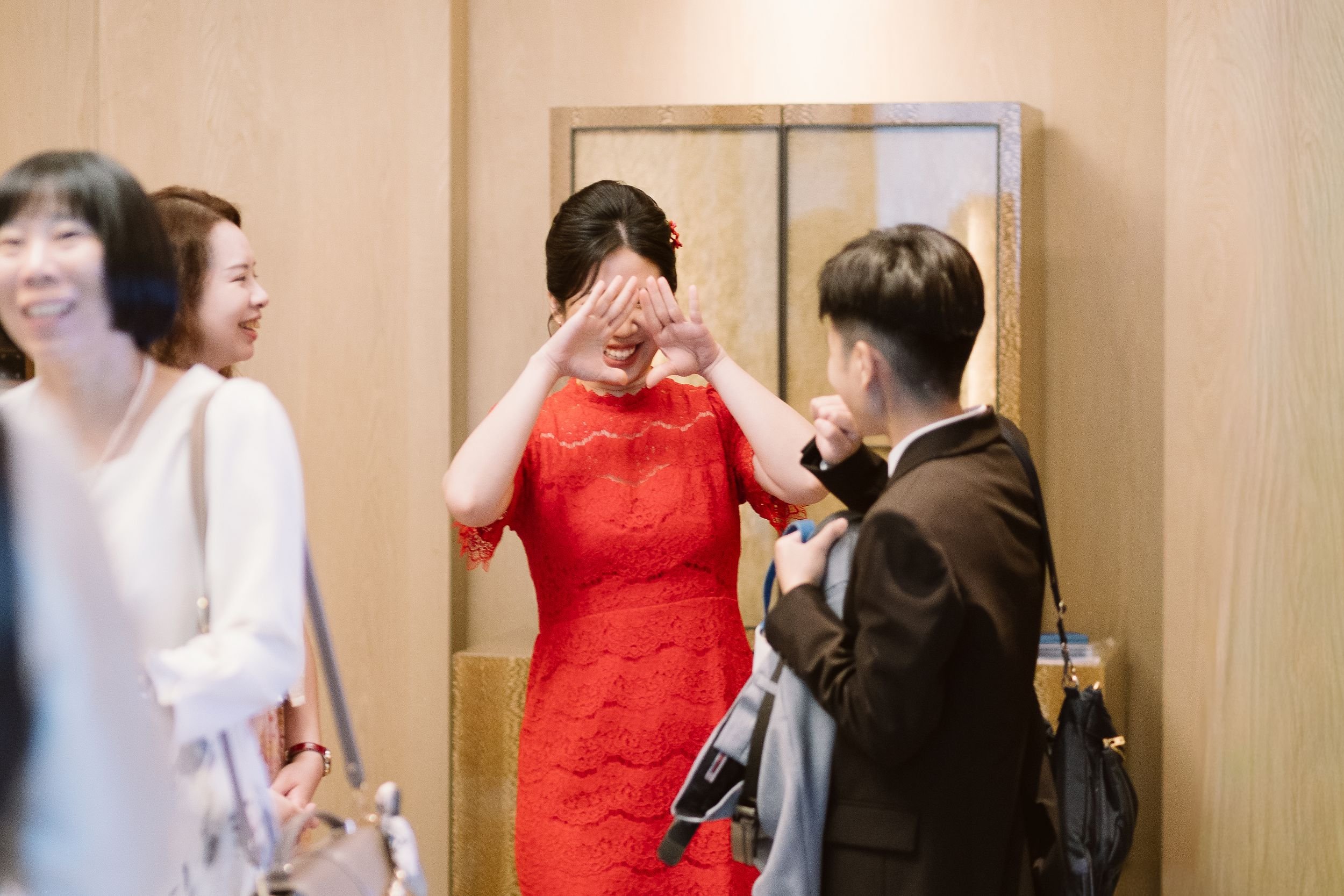A woman in a red lace dress is smiling and using her hands to form a triangle shape around her eyes while talking to a young man in a black jacket, who has a backpack. Two other women are partially visible on either side, smiling and listening. The b