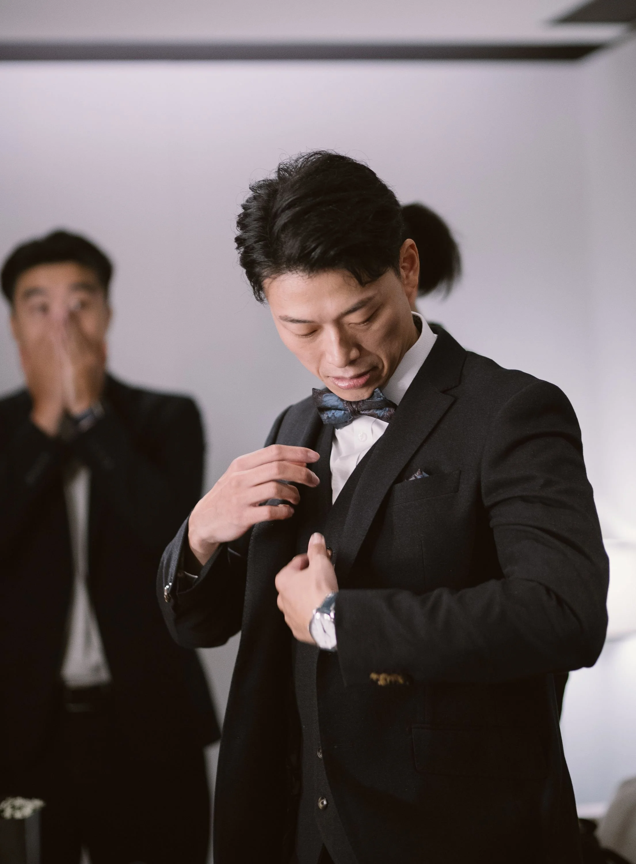 Man wearing a black tuxedo, adjusting his jacket, with another man in the background.