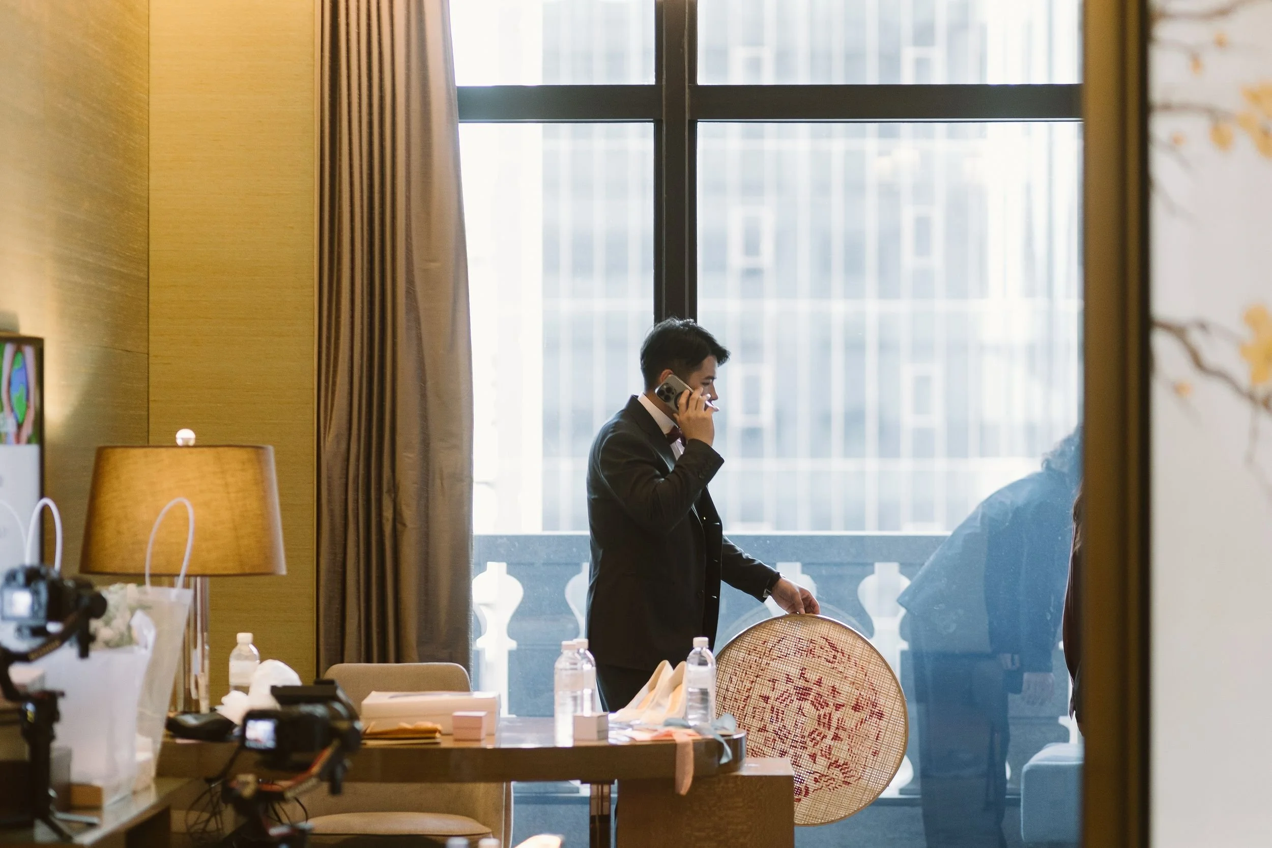 A man in a suit talking on a cell phone inside a room with a large window and beige curtains, with various items on a table in the foreground.