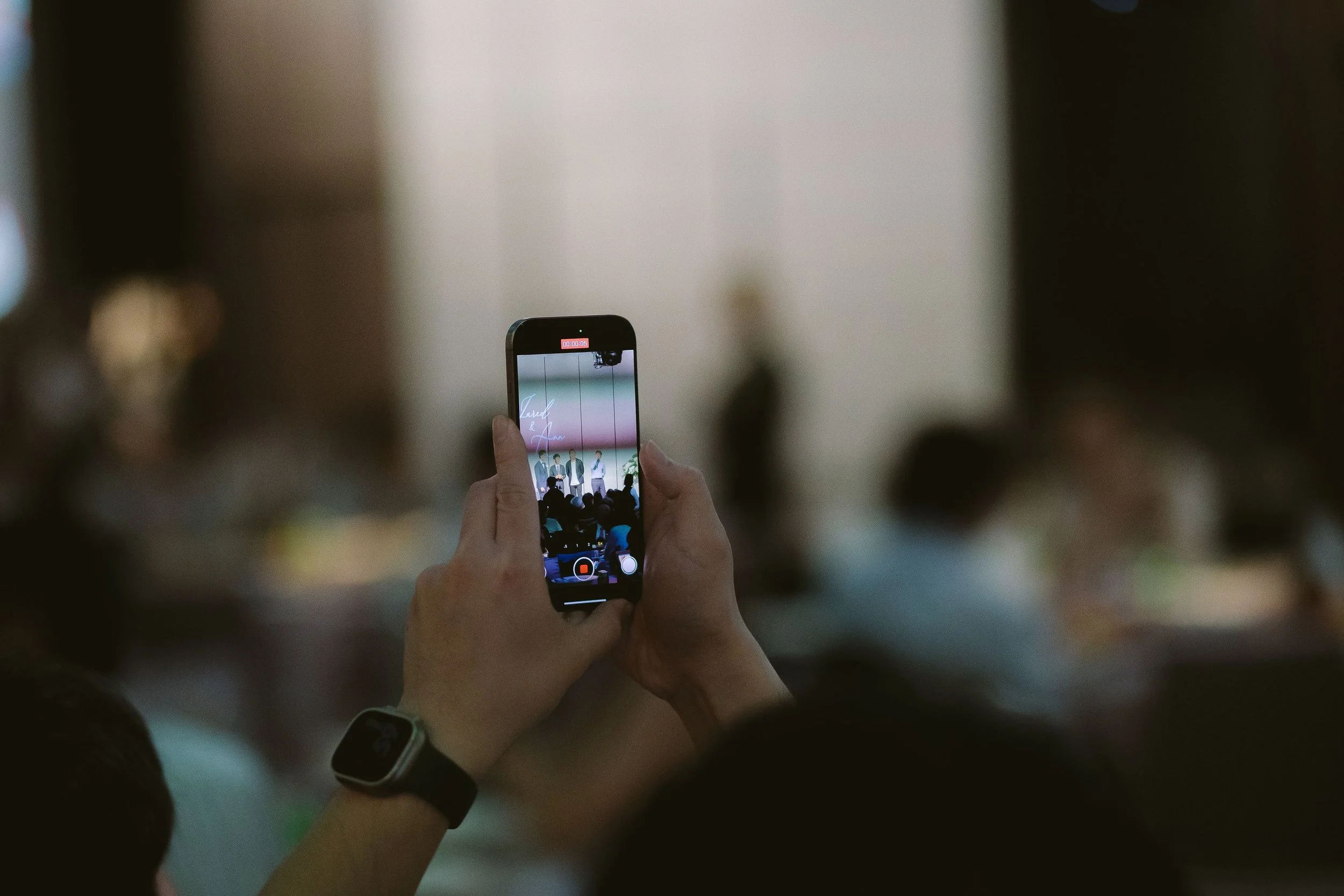 Person holding a smartphone recording a group of people on stage at an indoor event.