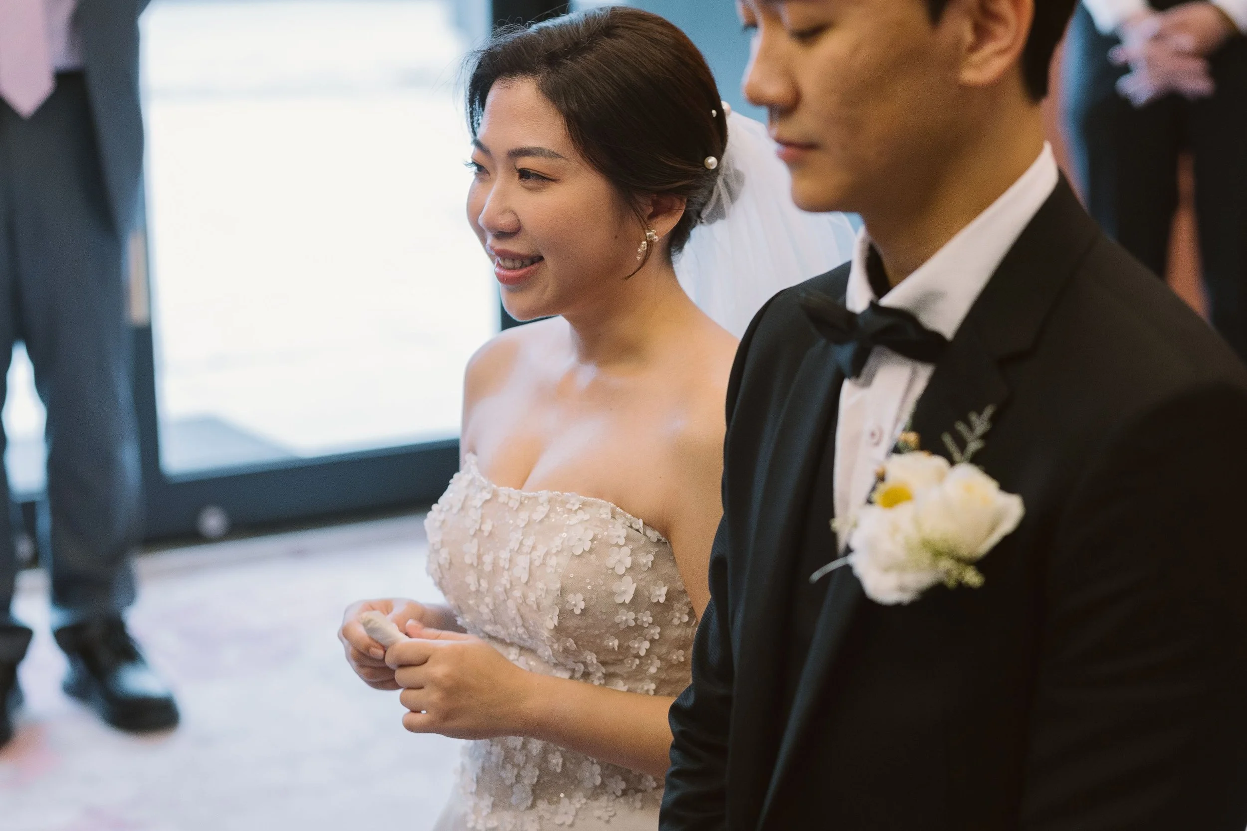 A bride and groom standing close together during a wedding ceremony, with the bride wearing a lace off-shoulder wedding gown and the groom in a black tuxedo with a bow tie and boutonniere, both smiling softly.