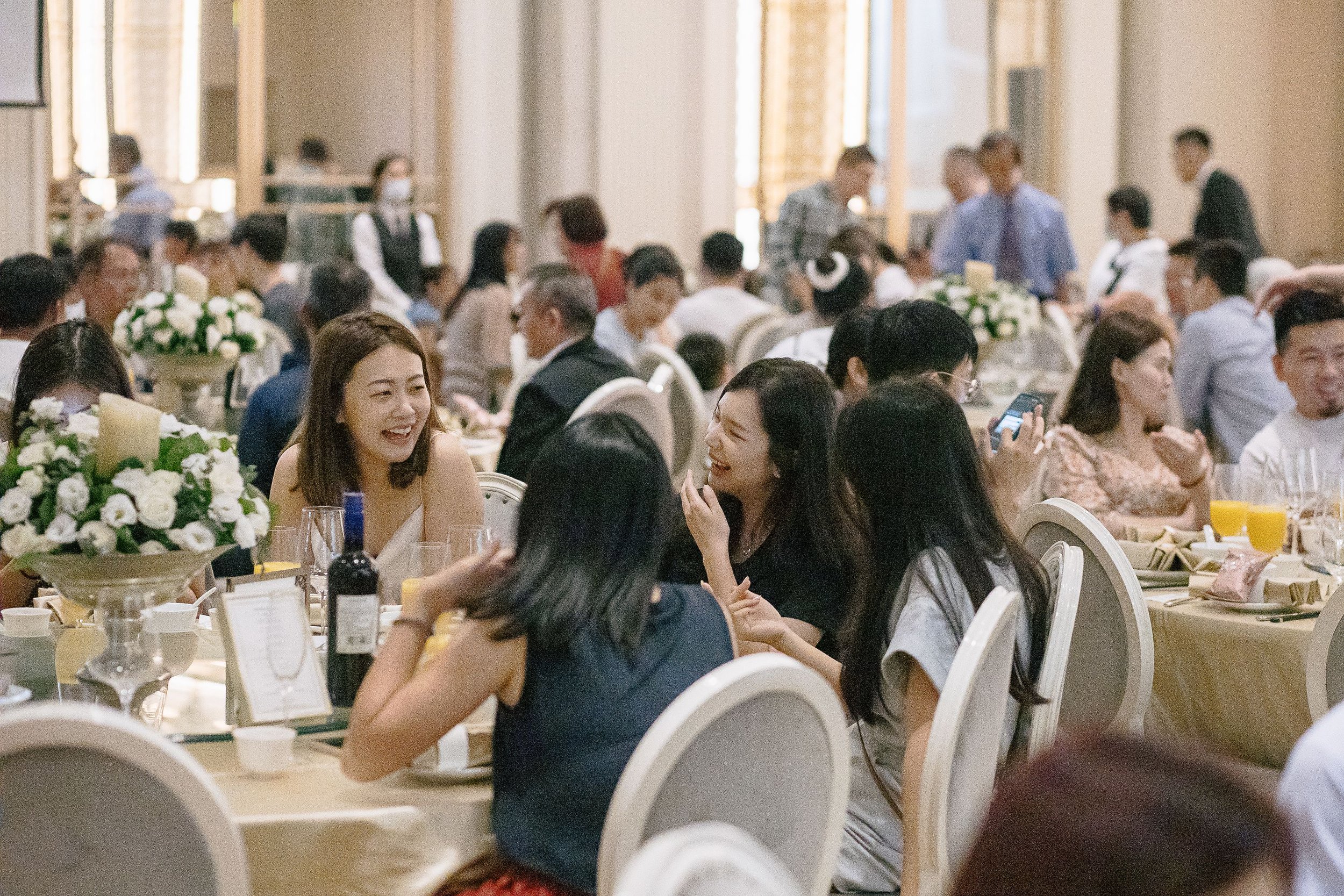 A busy banquet hall filled with people dressed in formal attire, sitting at round tables decorated with floral centerpieces and candles, enjoying a social gathering or celebration.
