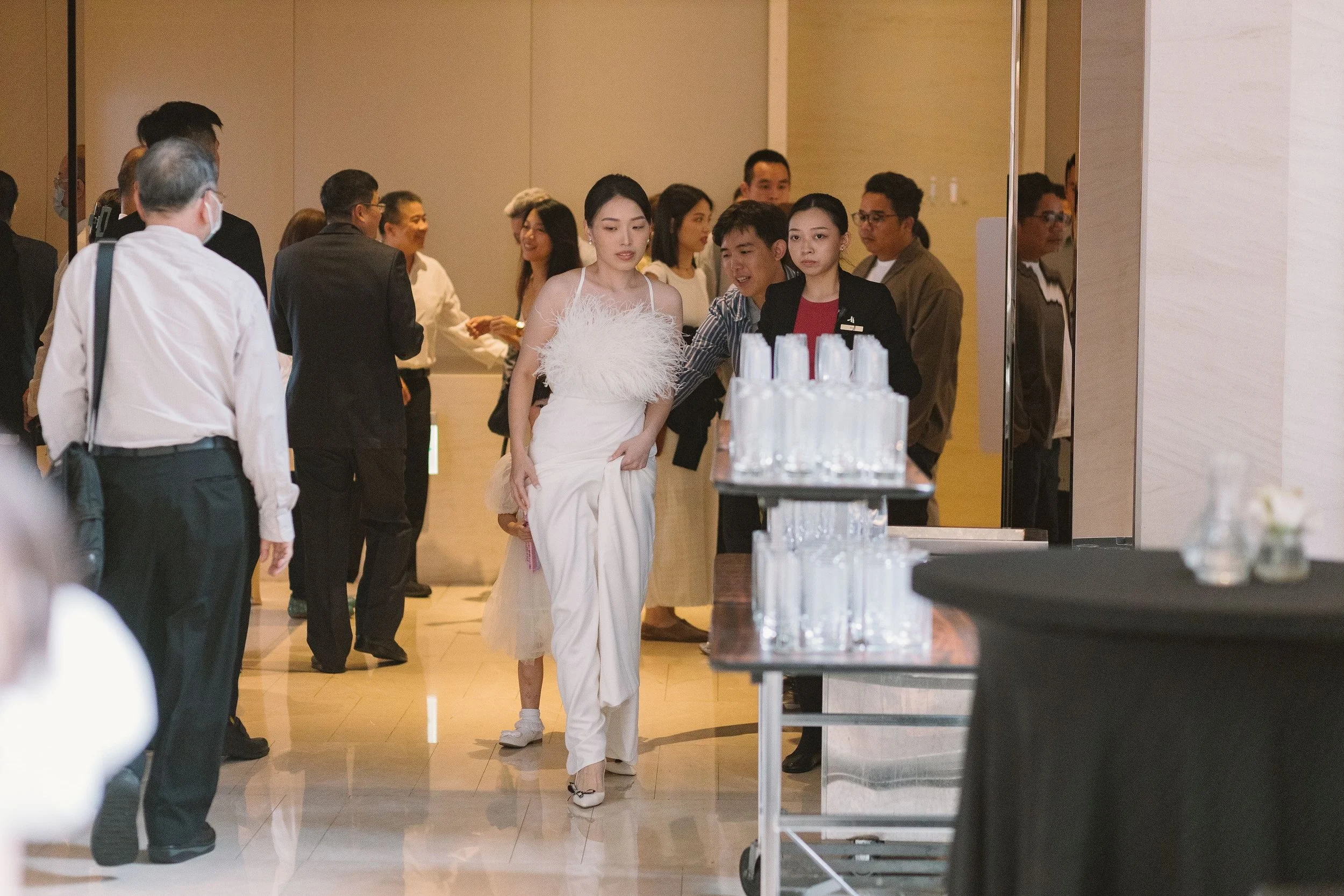 A woman in a white dress walking through a crowded indoor space, possibly a hotel or conference center, with group of people around her.