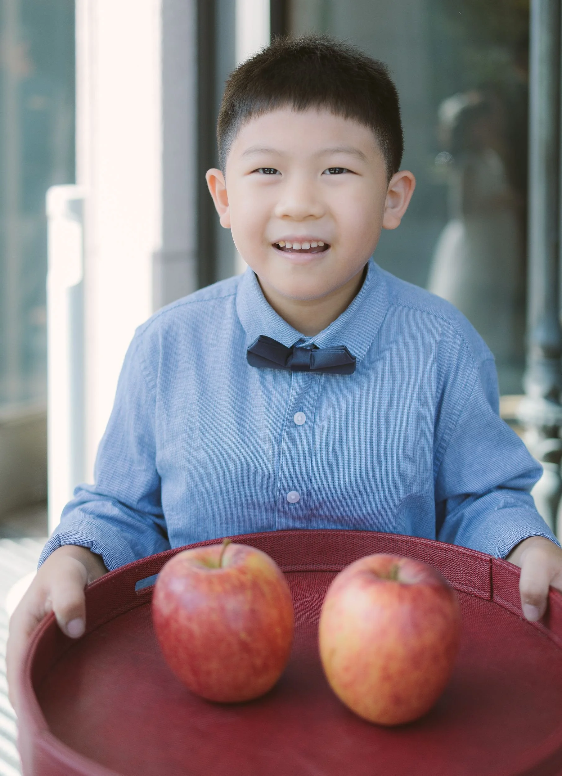 A young boy with short black hair and a blue shirt with a black bow tie smiling at the camera while holding a red basket filled with red apples.
