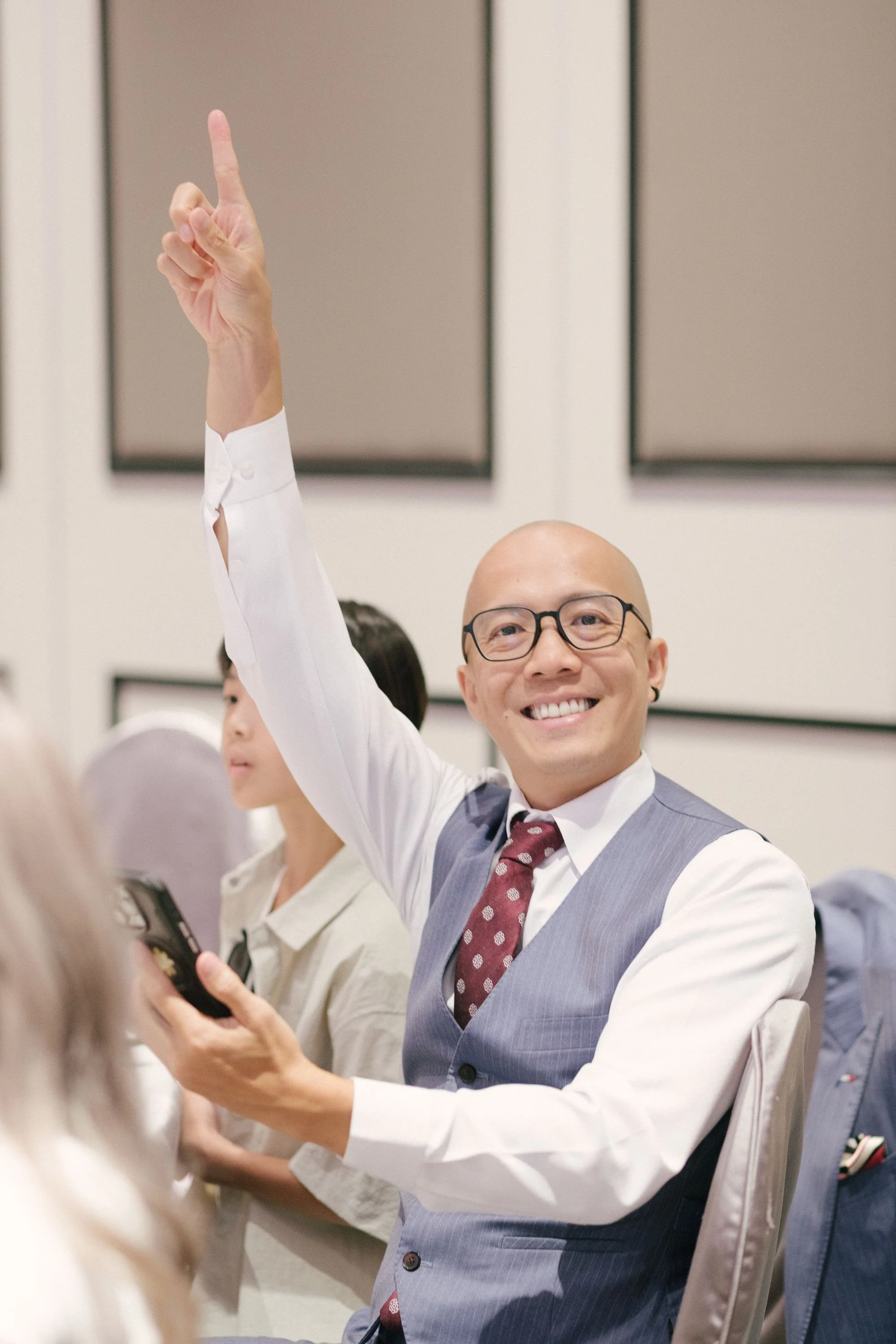 A man in a gray vest and dress shirt with a maroon polka dot tie, wearing glasses, is smiling and raising his hand with his index finger pointing up during a meeting or conference.