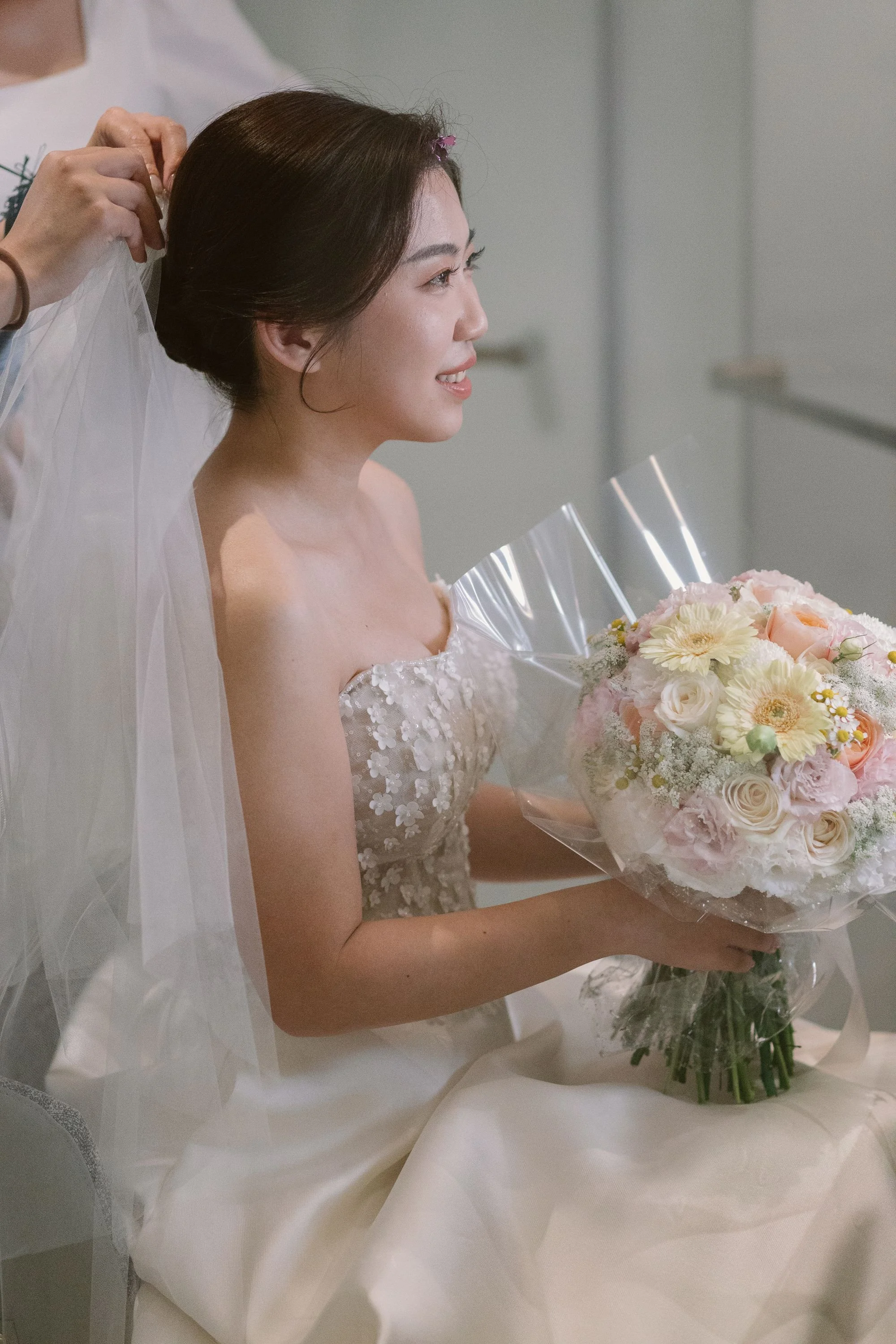 A bride holding a bouquet of pink and white flowers, smiling, with a veil being adjusted on her head, wearing an off-the-shoulder wedding dress with floral embellishments.