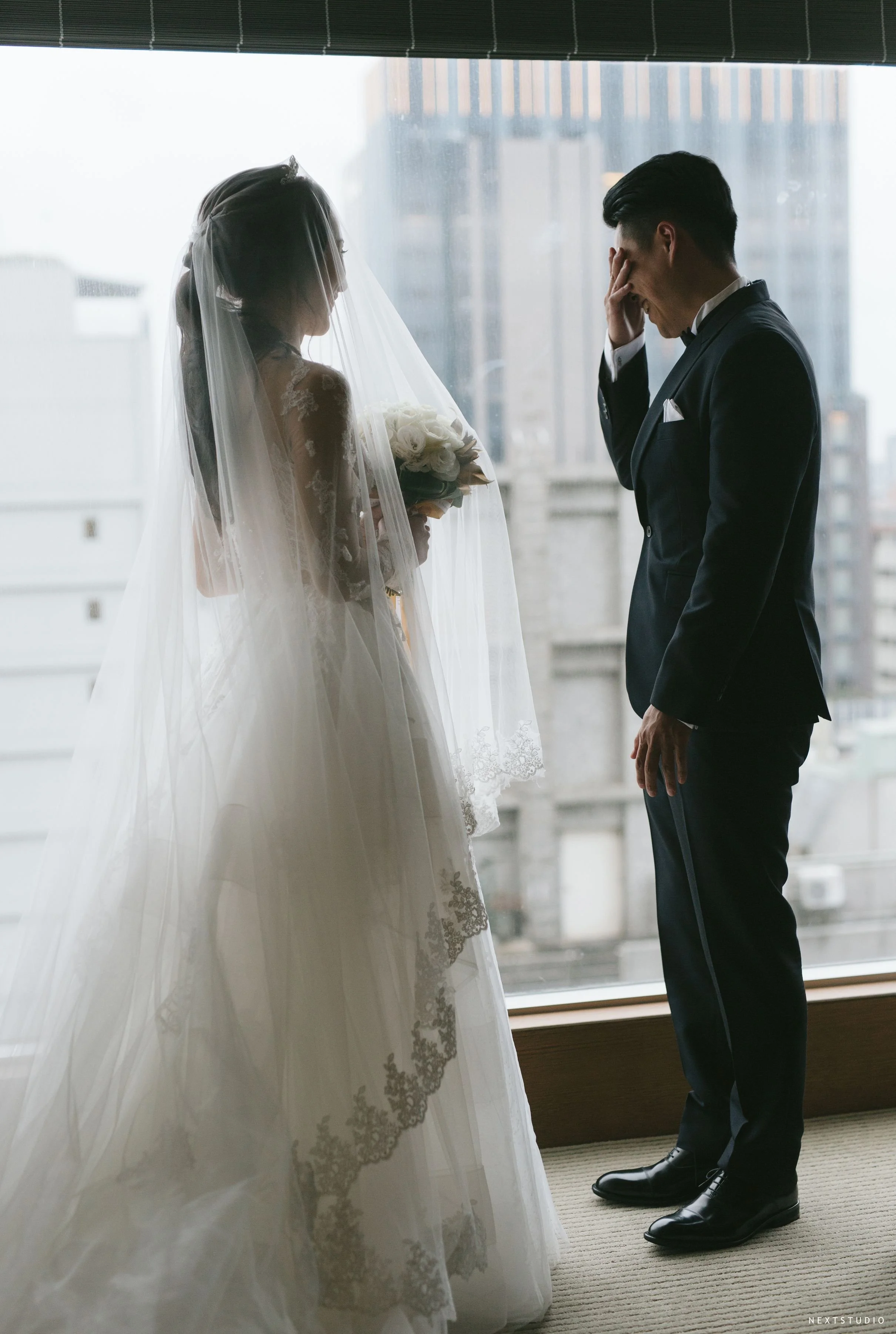 A bride in a wedding gown holding a bouquet of white roses, standing in front of a window, smiling at a groom dressed in a black suit who is covering part of his face with his hand, smiling back.
