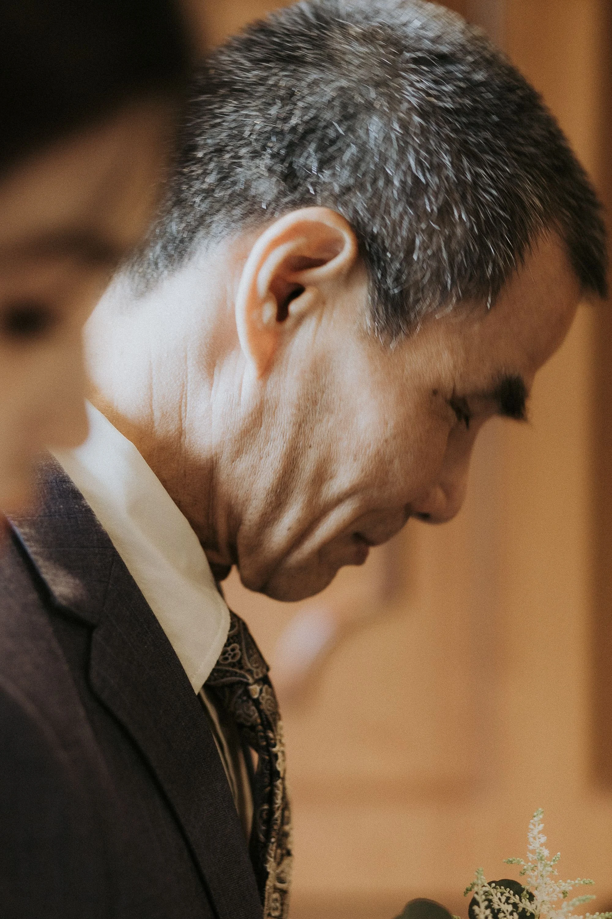 Close-up of a man with graying hair wearing a dark suit, white shirt, and patterned tie, focusing on his face and upper body, with some flowers in the foreground.