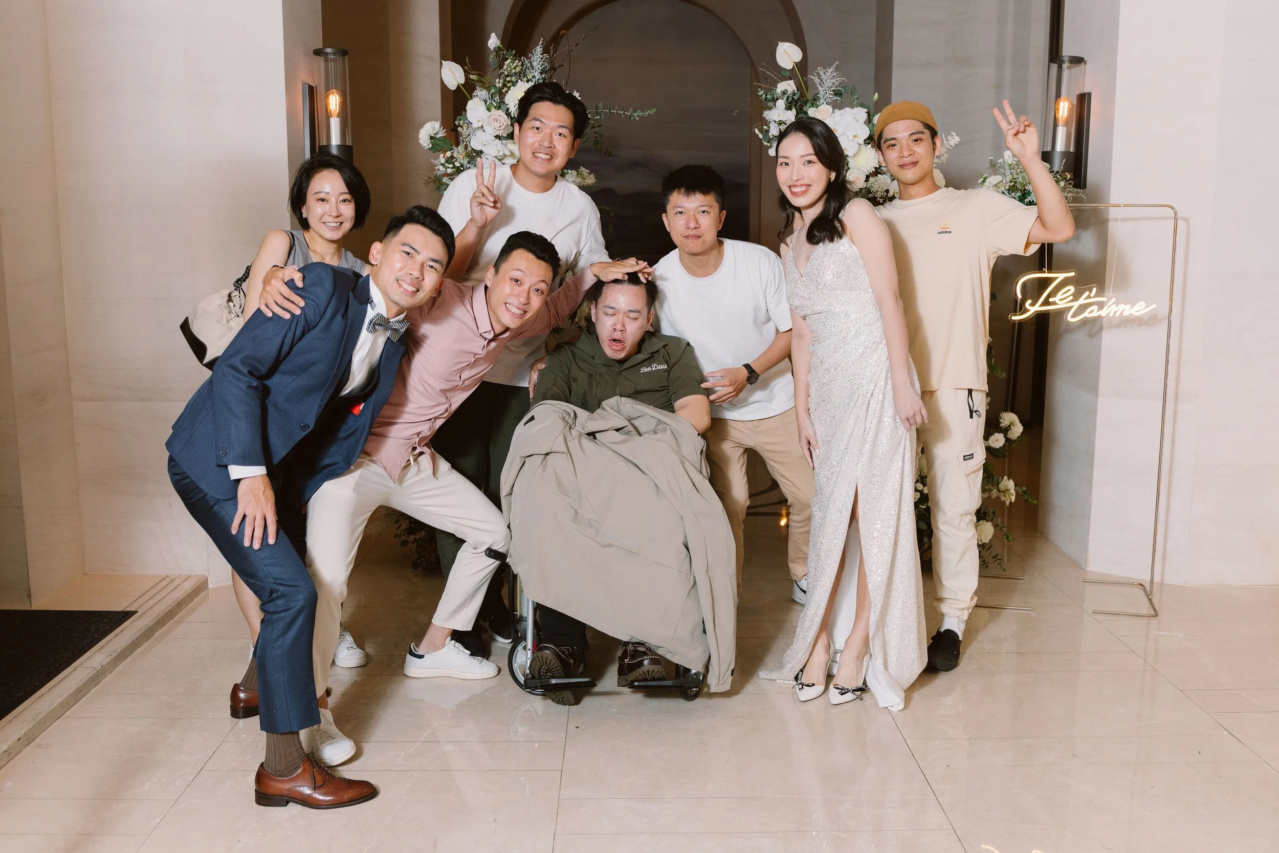 Group of seven people, five women, and two men, smiling and posing at an indoor event. One woman is sitting in a wheelchair at the center. Background features floral arrangements and a sign that reads 'Je t'aime'.
