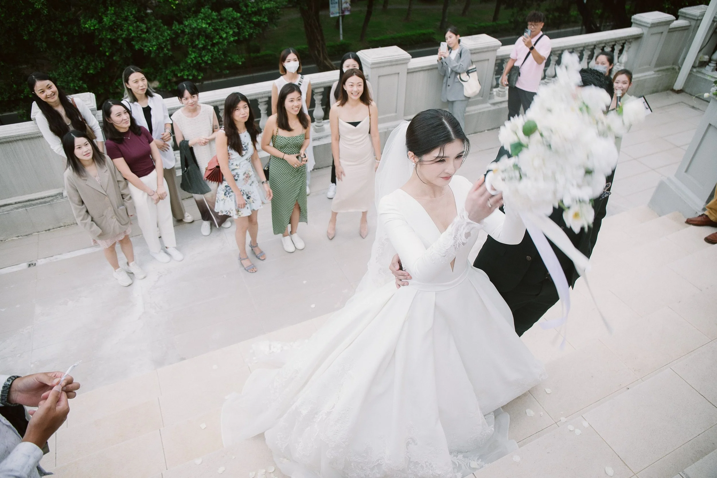 A bride in a white wedding dress holding a bouquet of white flowers during her wedding ceremony, surrounded by friends and family taking photos and videos on their phones. The scene takes place outdoors on the steps of a building with a decorative ra