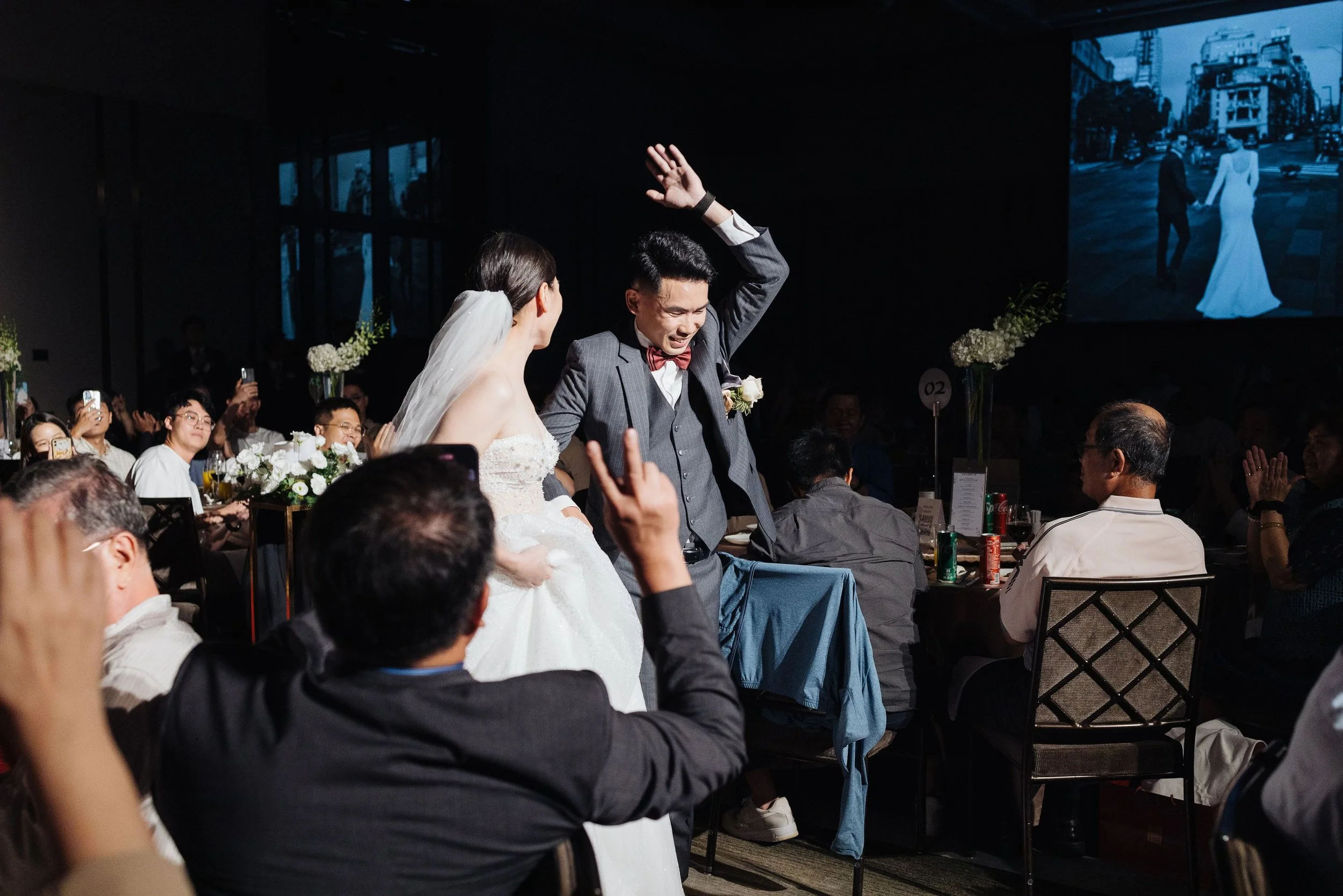 A wedding reception with the bride and groom dancing in the spotlight, surrounded by guests seated at tables, with a large screen displaying a photo of a couple holding hands in the background.
