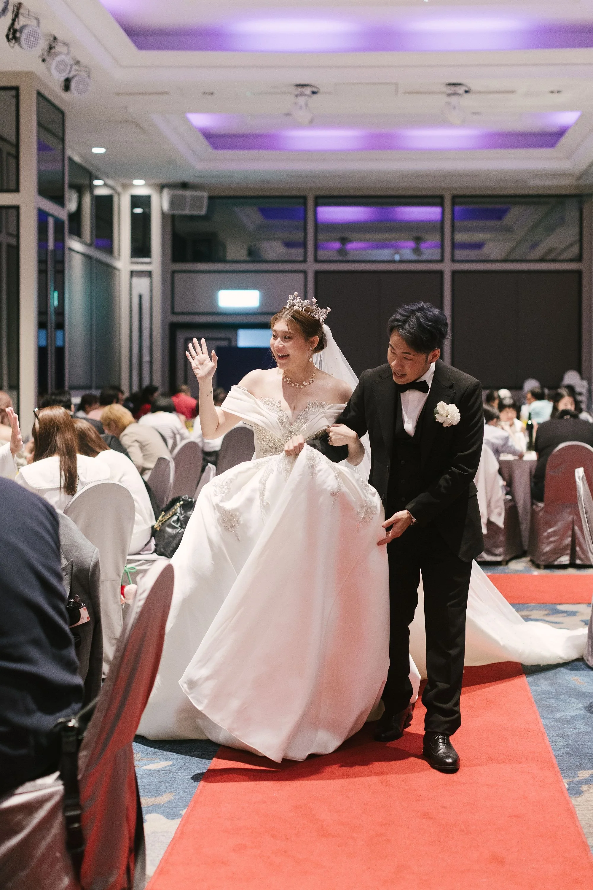 A bride and groom dancing during their wedding reception inside a banquet hall with purple ceiling lighting, surrounded by seated guests.