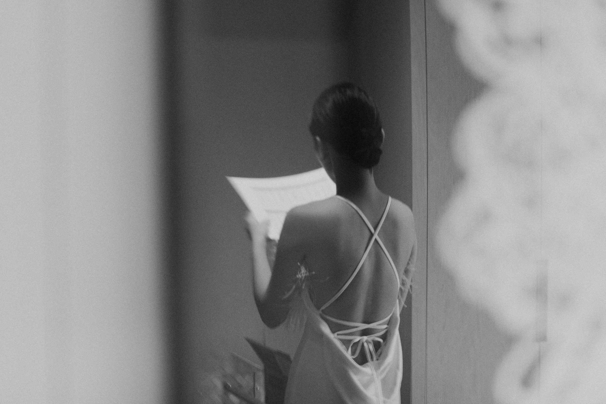 A woman standing in front of a mirror, seen from behind. She is dressed in a swimsuit or lingerie with crisscross straps at the back. The photo is black and white, capturing her reflection and hairstyle.