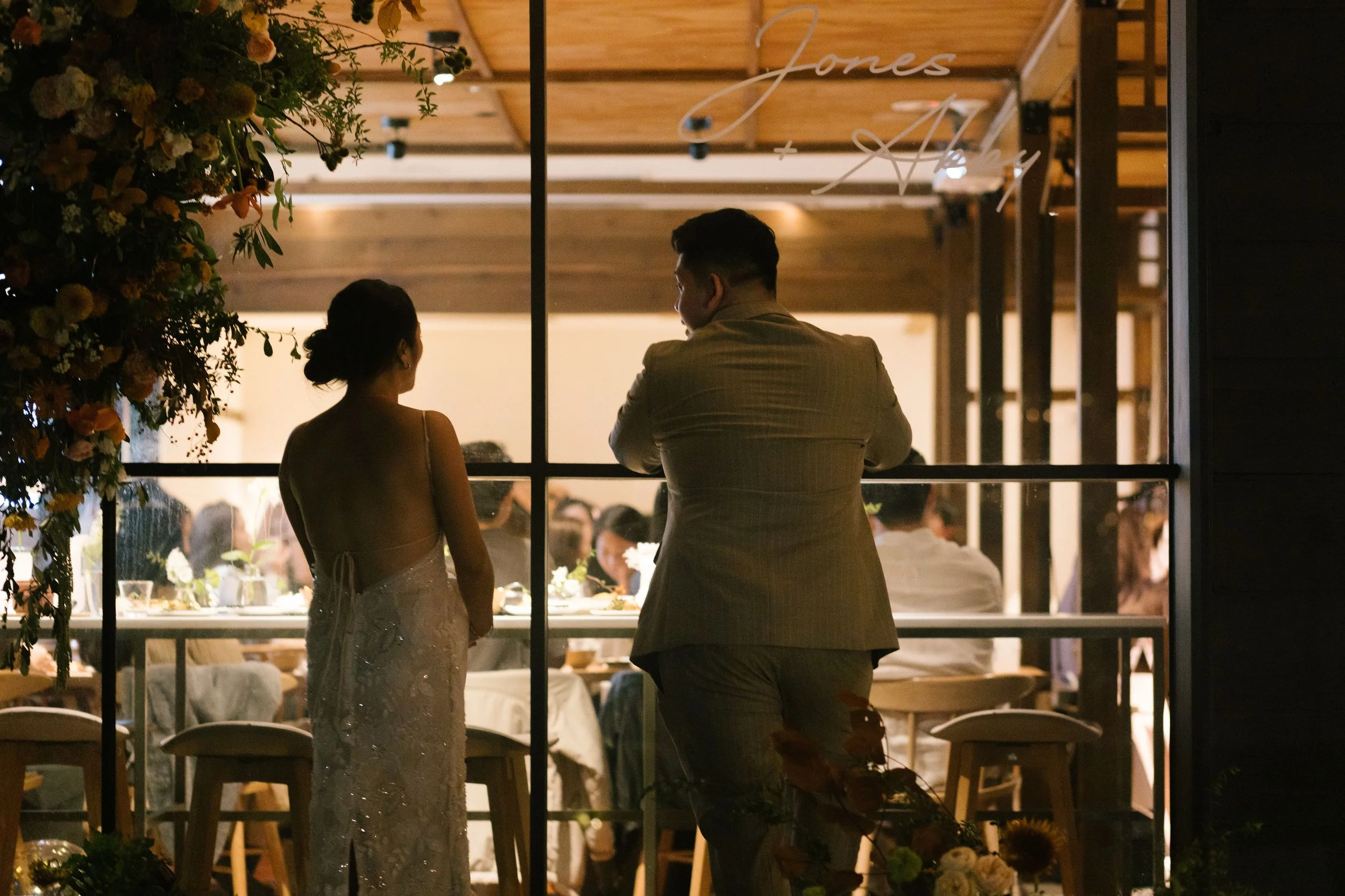 A bride and groom standing and talking in a room filled with guests, seen through a window at night during a wedding celebration.