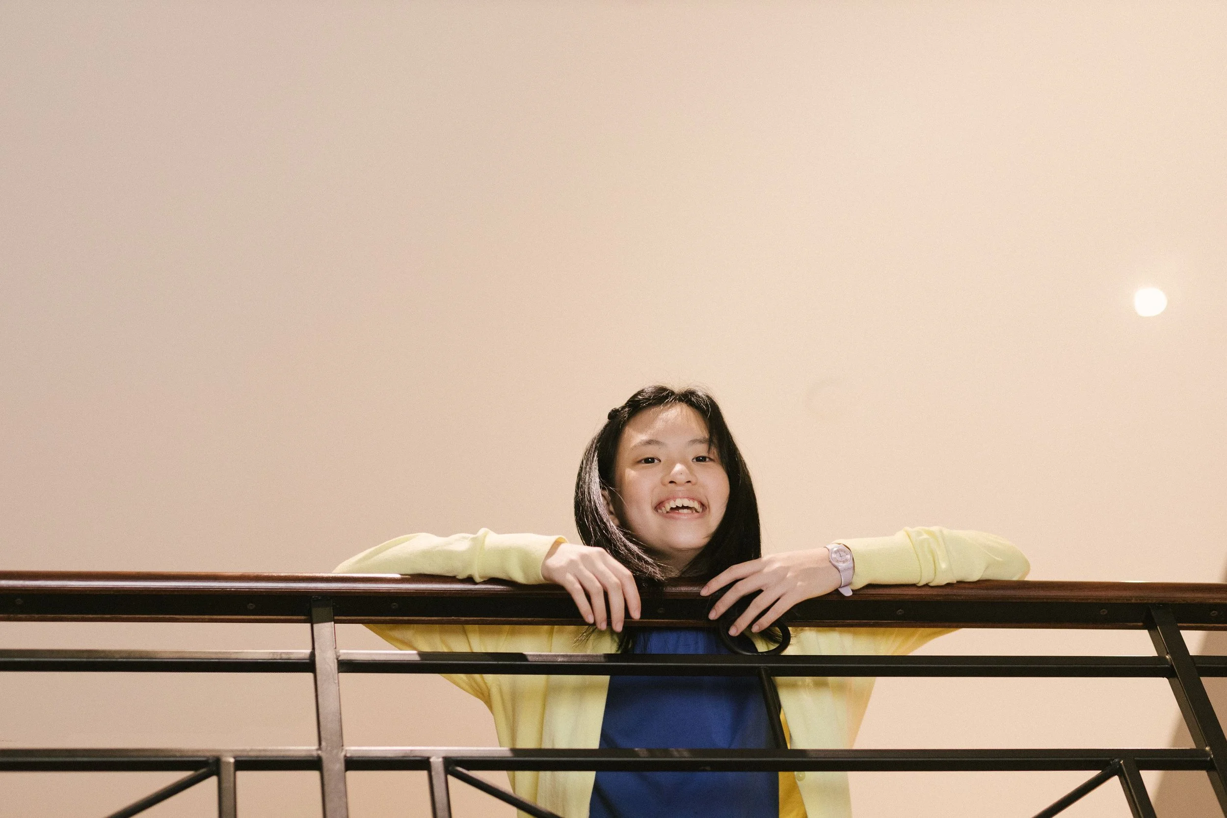 A young girl with shoulder-length black hair smiling and leaning over a railing in an indoor setting, wearing a yellow jacket and a blue shirt.
