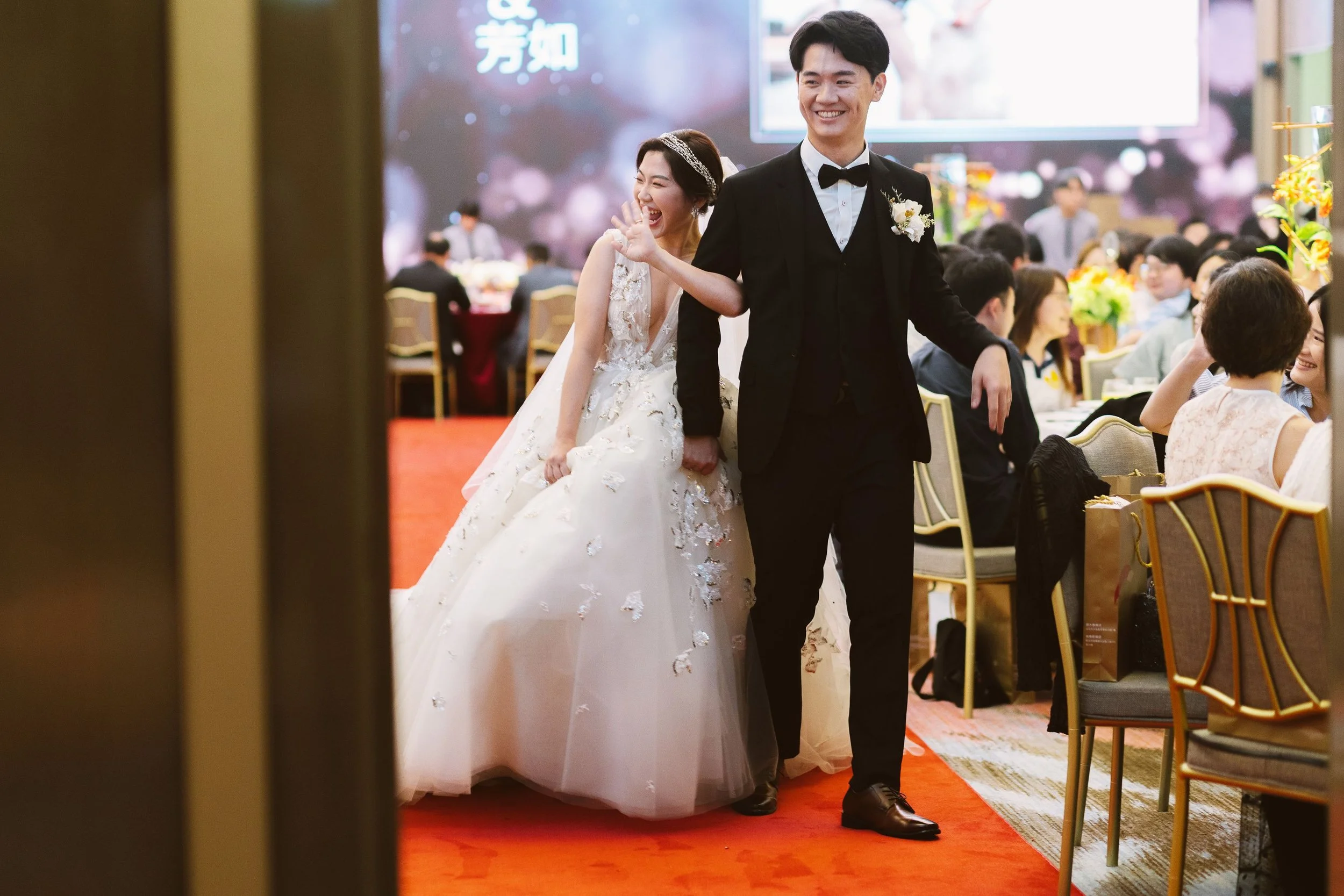 A bride with a wedding dress and a groom in a tuxedo walking through a banquet hall, smiling and celebrating during a wedding reception.