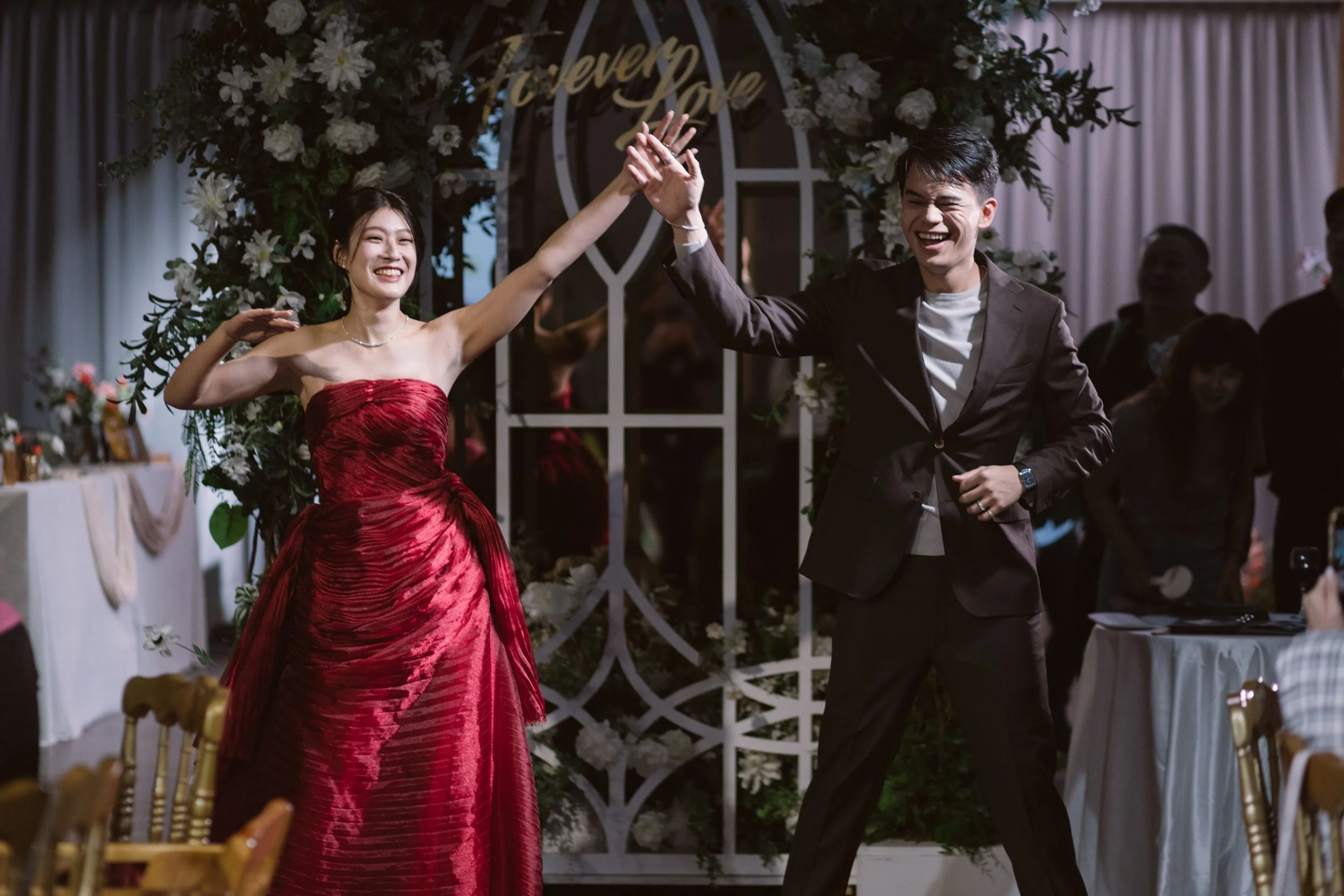 A young couple dancing and smiling at a wedding reception. The woman is wearing a strapless red dress, and the man is wearing a dark suit with a white shirt. They are holding hands and appear happy. The background has floral decorations and a sign th