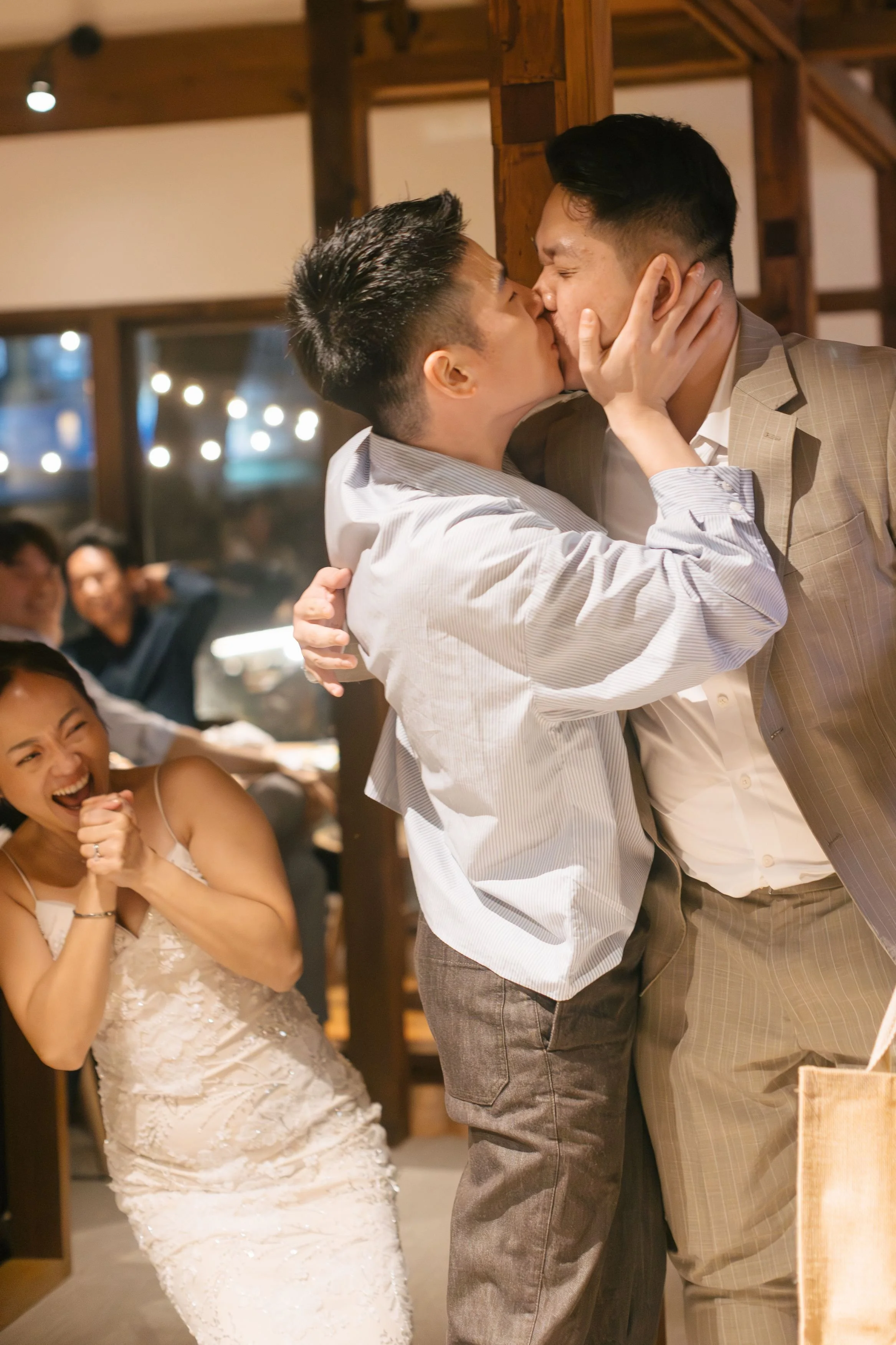 Two men are kissing while a woman in a white dress laughs and covers her mouth with her hand.