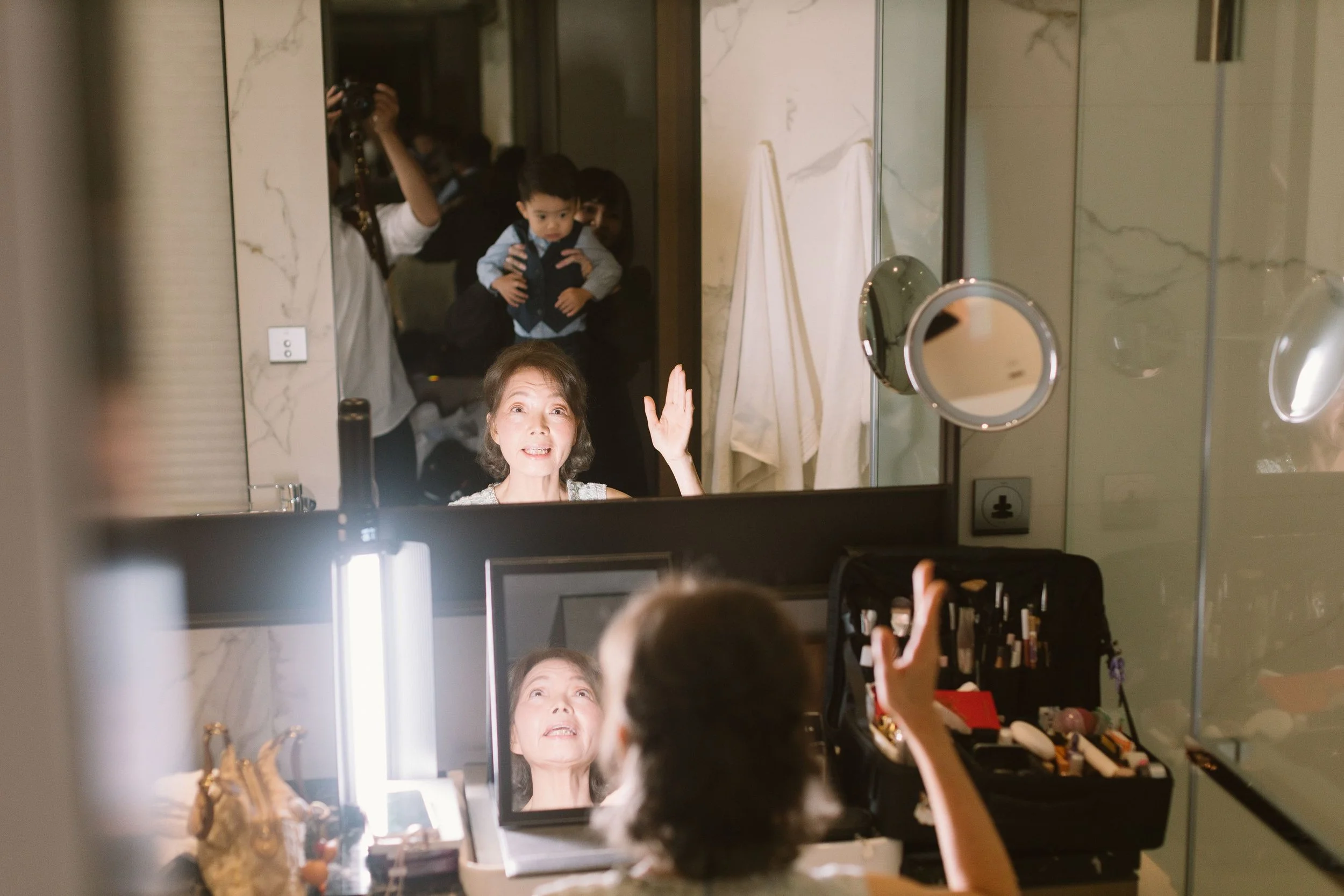 A woman with short gray hair is looking into a mirror, applying makeup or lipstick, while a young boy, possibly her grandson, stands beside her in front of the mirror, and a person behind them holds a camera capturing the scene. The room has a marble