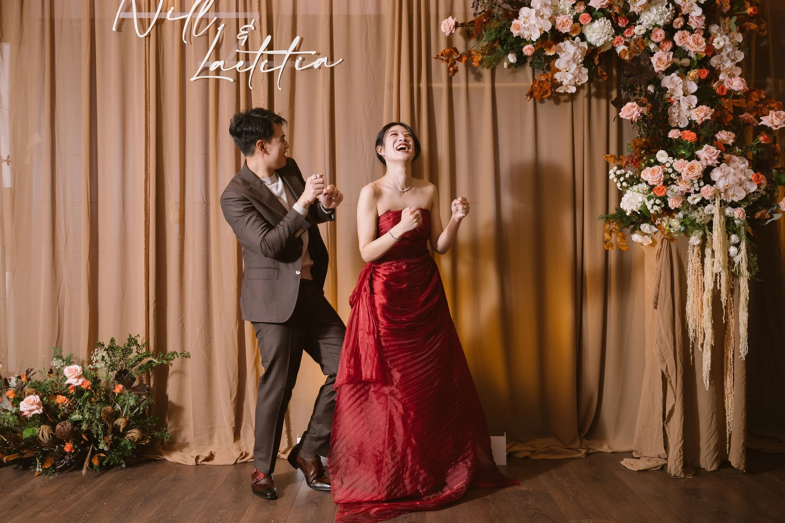 A joyful couple dancing at a celebration with floral decorations and a beige curtain backdrop.