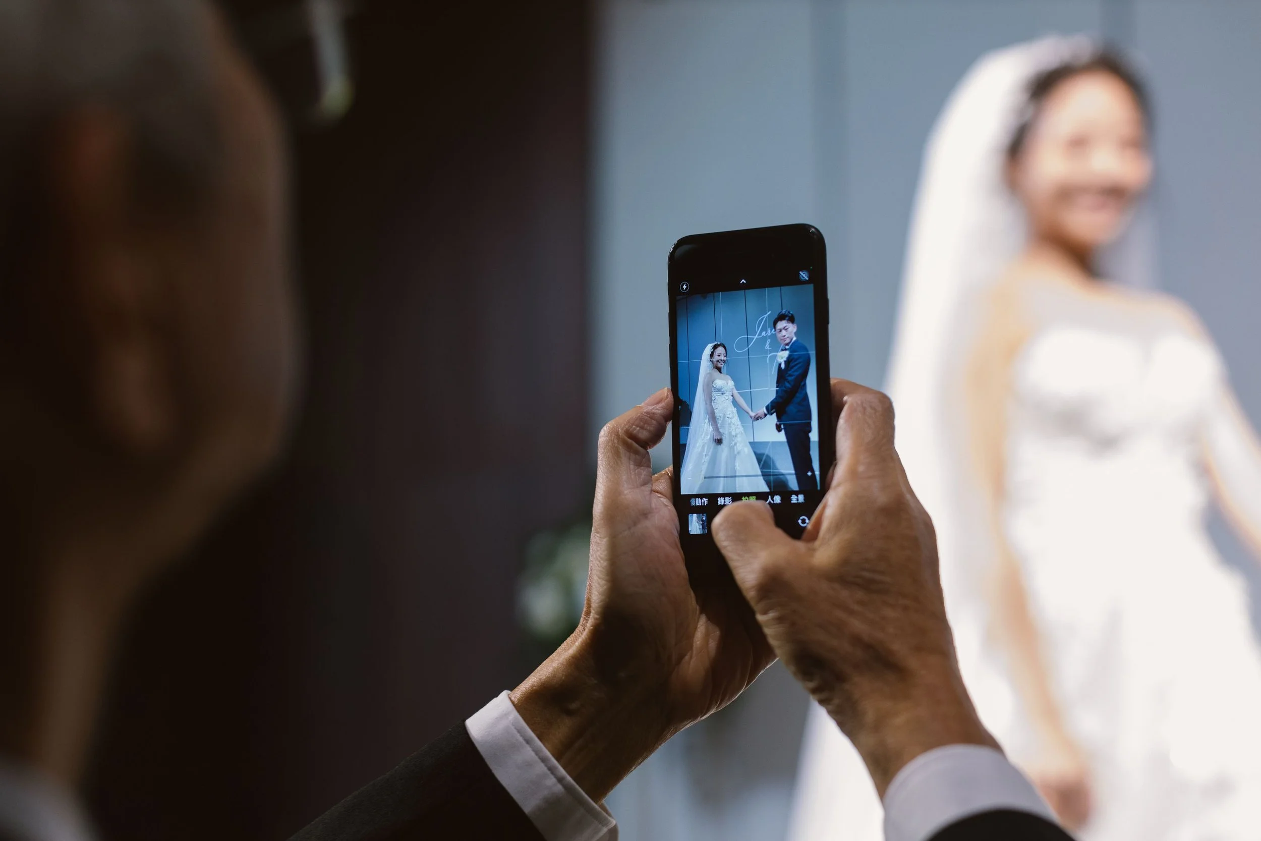 Person holding a smartphone taking a photo of a bride and groom on a wedding day.